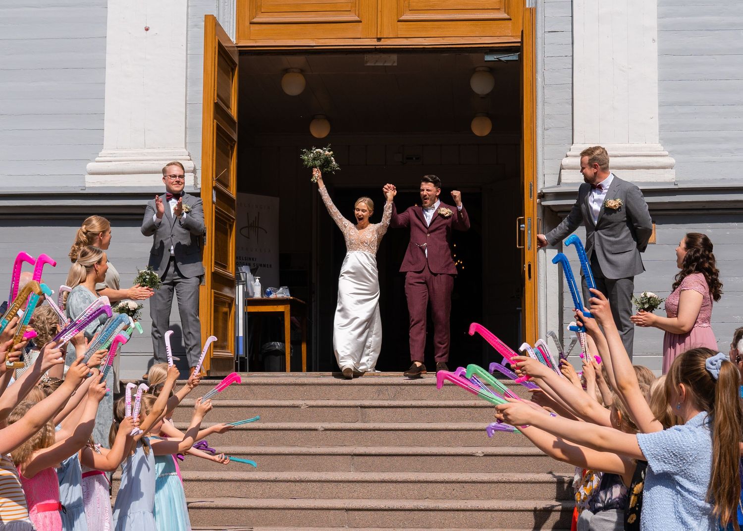 Newlyweds celebrate on church steps raising bouquet and fist while guests wave colorful streamers.