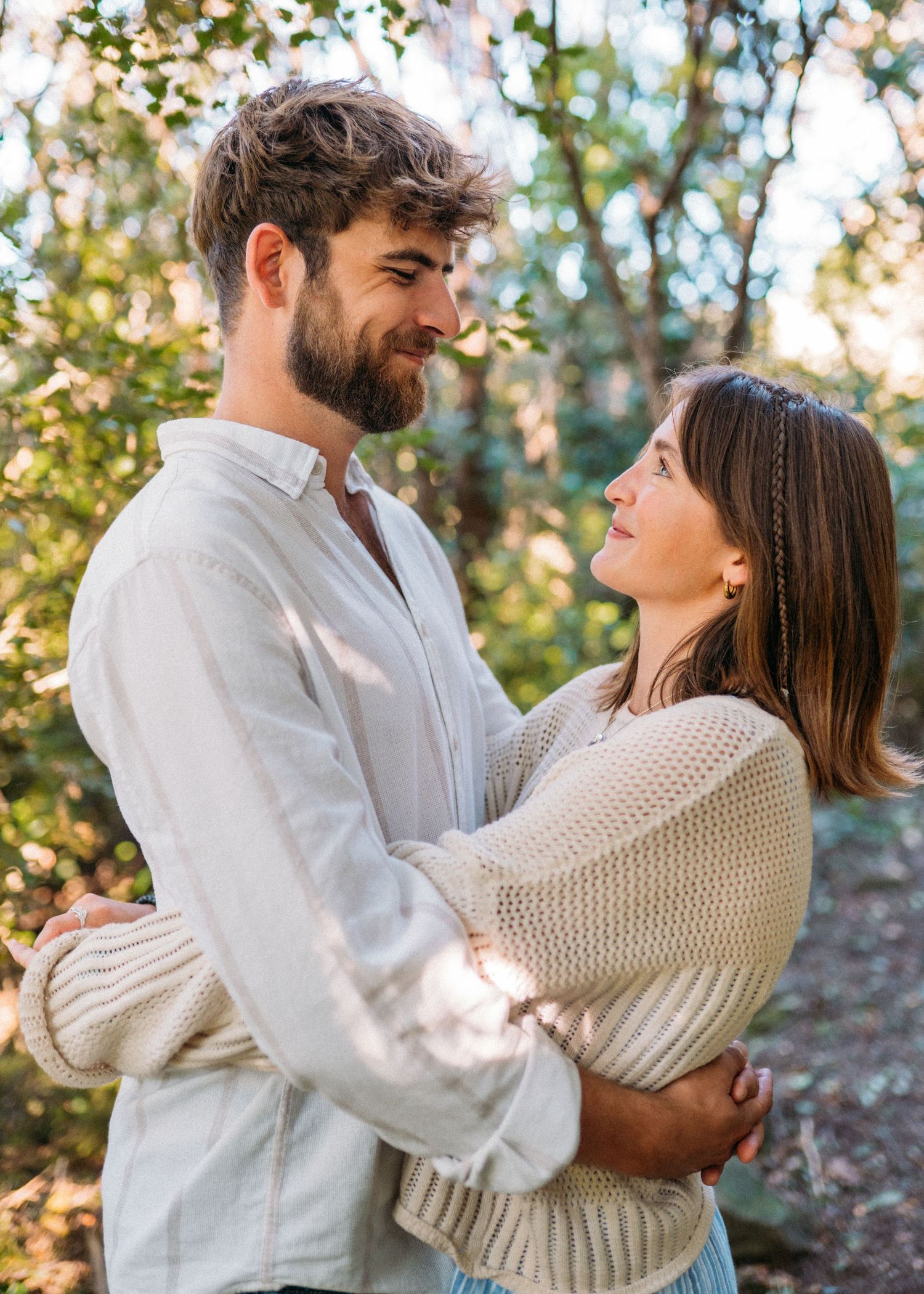 Couple holding each other close and smiling in a bright sunlit woodland setting outdoors.