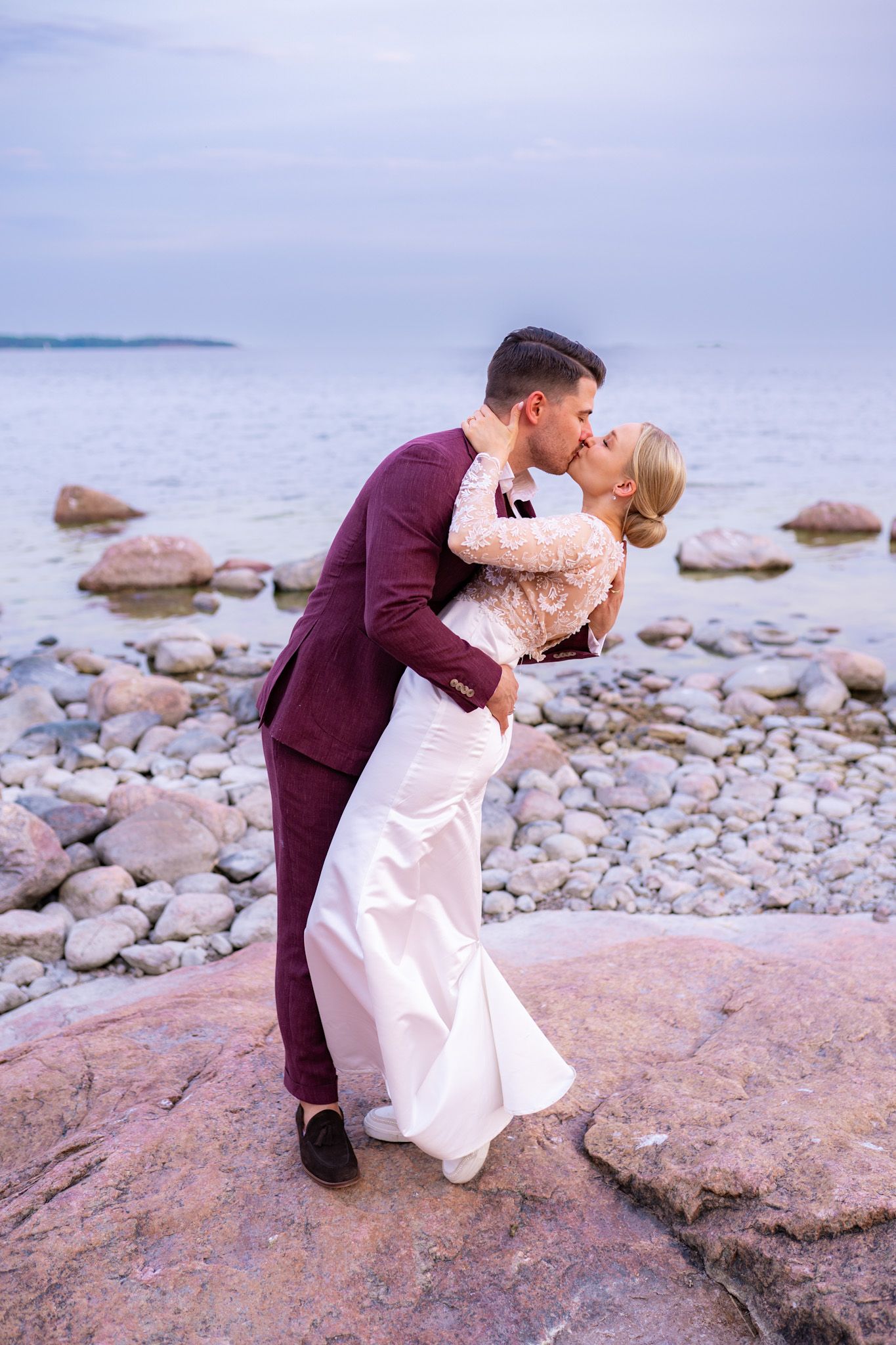 Bride and groom sharing a romantic dip kiss on a rocky shoreline at dusk with calm sea in background.