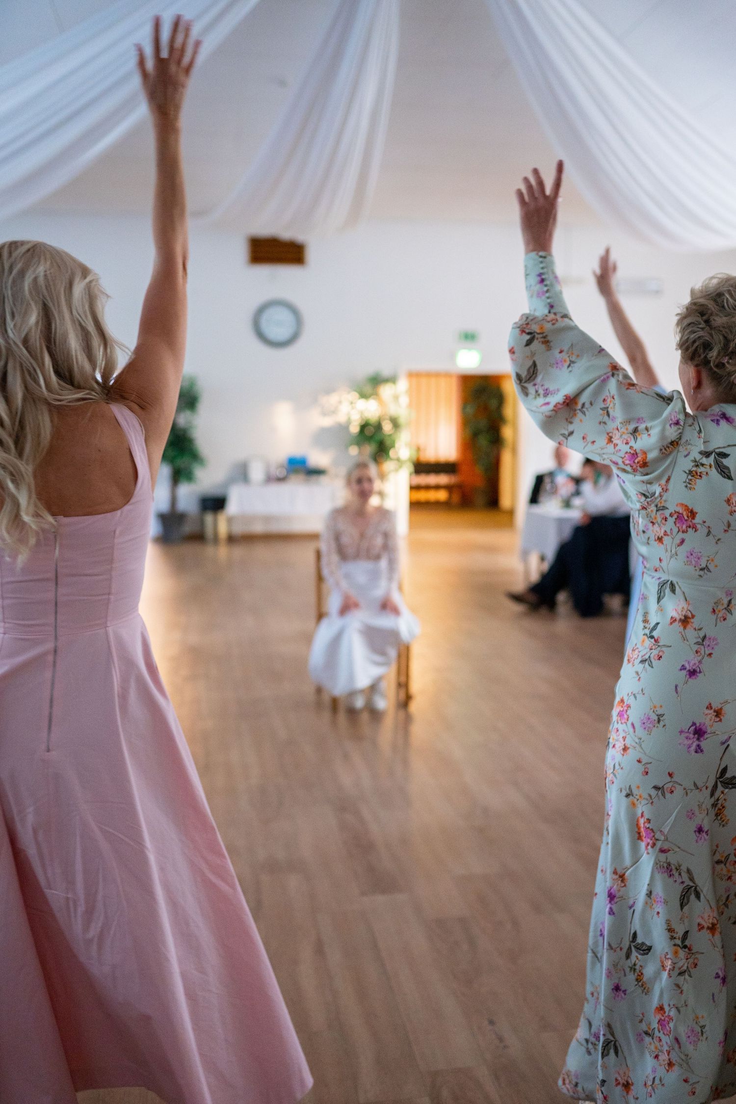 Two women raising arms toward seated bride during surprise wedding reception dance performance.