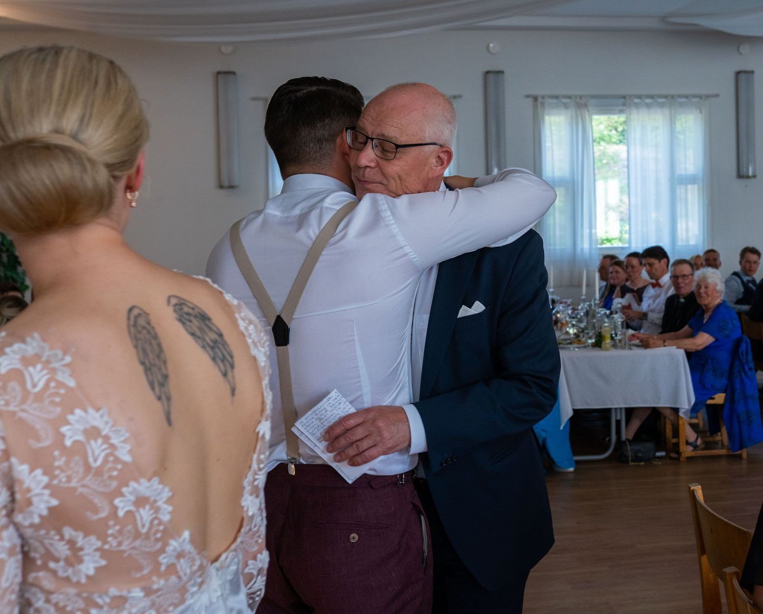 Groom in burgundy suit embracing an older man while bride watches at wedding reception.