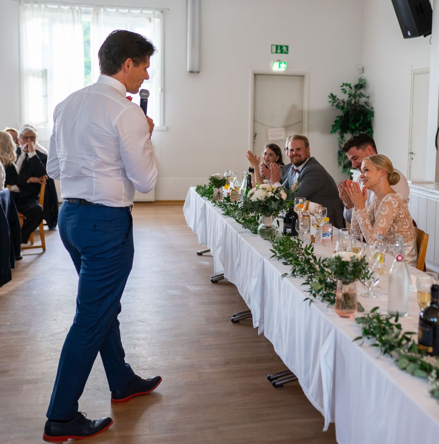 Best man gives a speech to laughing wedding party seated at a decorated head table with floral garlands.