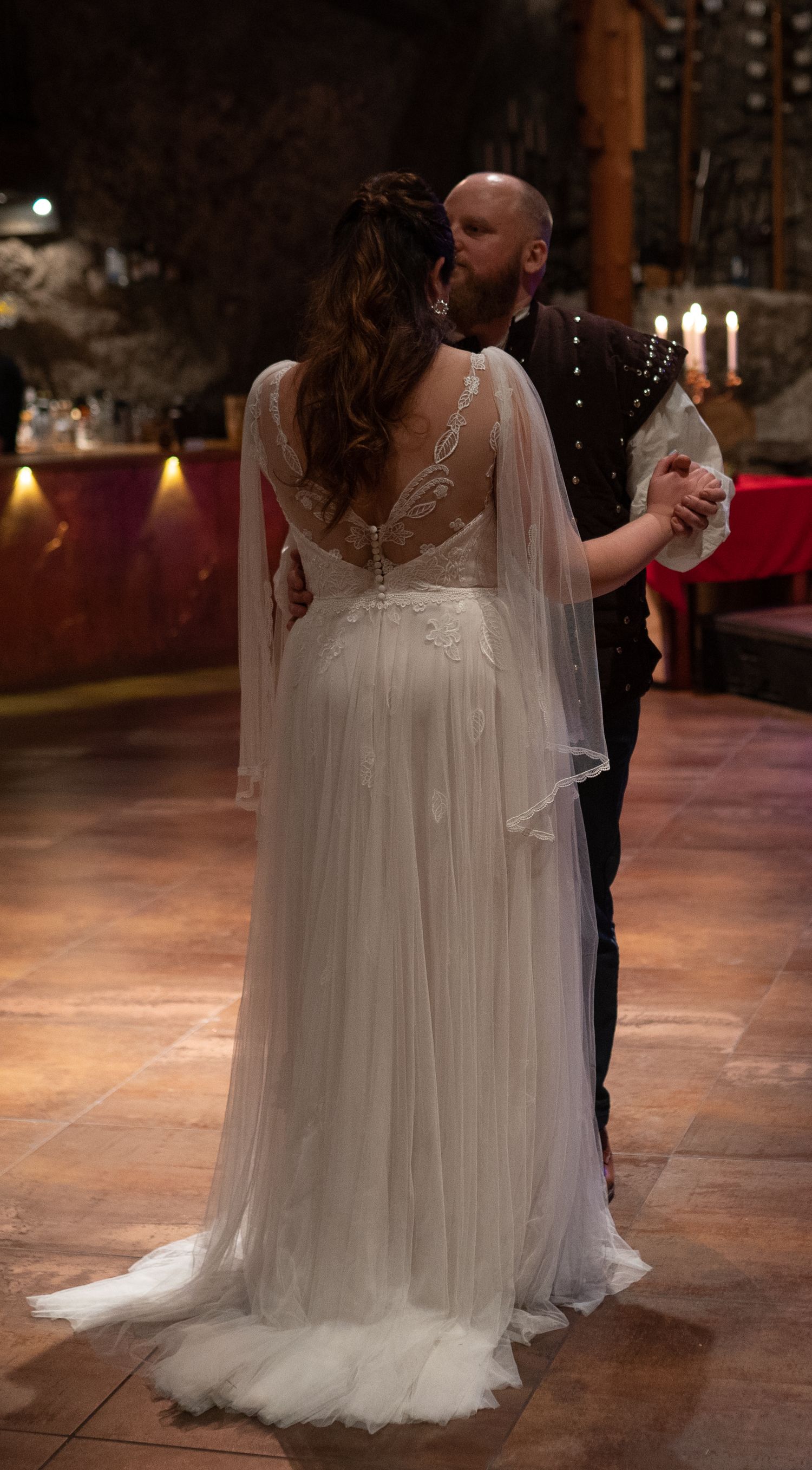 A bride in a flowing white lace wedding gown with cape sleeves shares a first dance with her groom in a warmly lit venue.