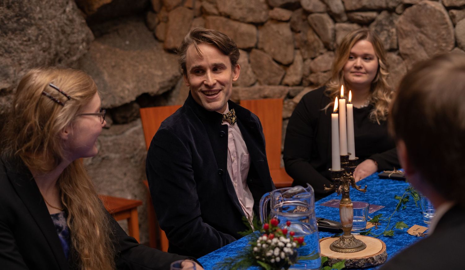 People smiling and talking at a candlelit dinner table with blue tablecloth and stone wall backdrop.