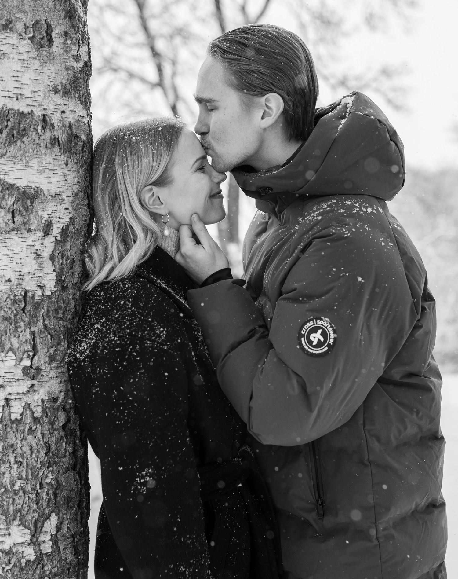 Black and white photo of a couple kissing near a birch tree in falling snow, wearing winter coats outdoors.