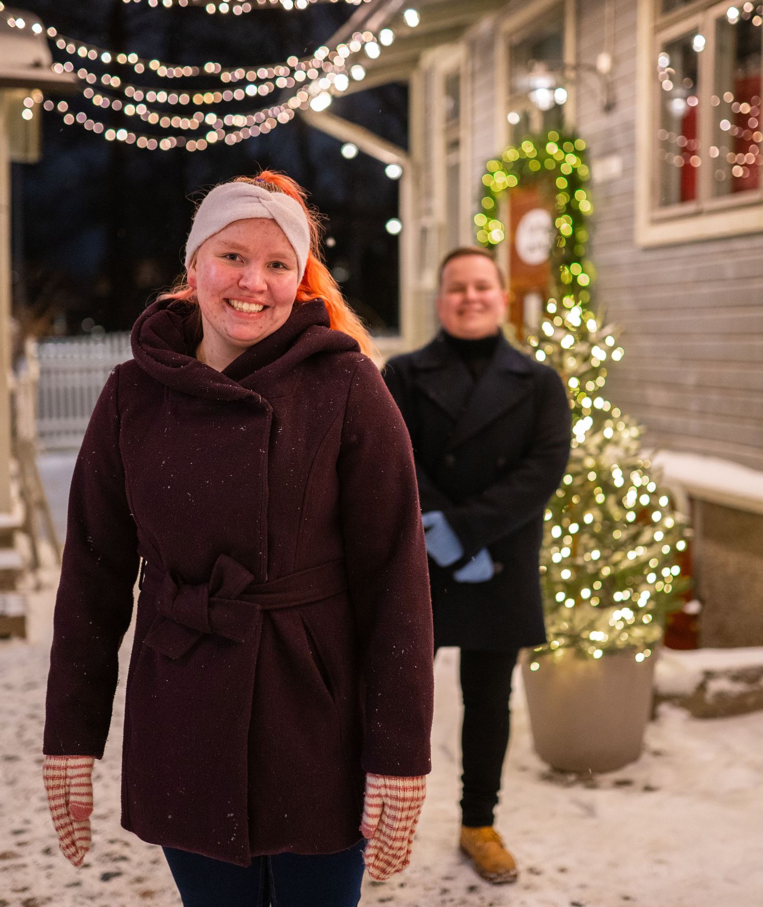 Two people smiling outdoors in winter, surrounded by festive Christmas lights and a decorated tree in a snowy setting.