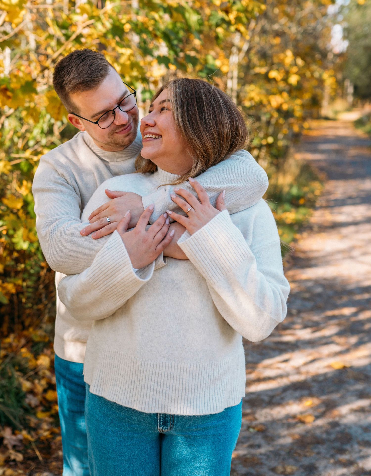Couple embracing on autumn forest path, woman smiling joyfully in cream sweater, man hugging her from behind.
