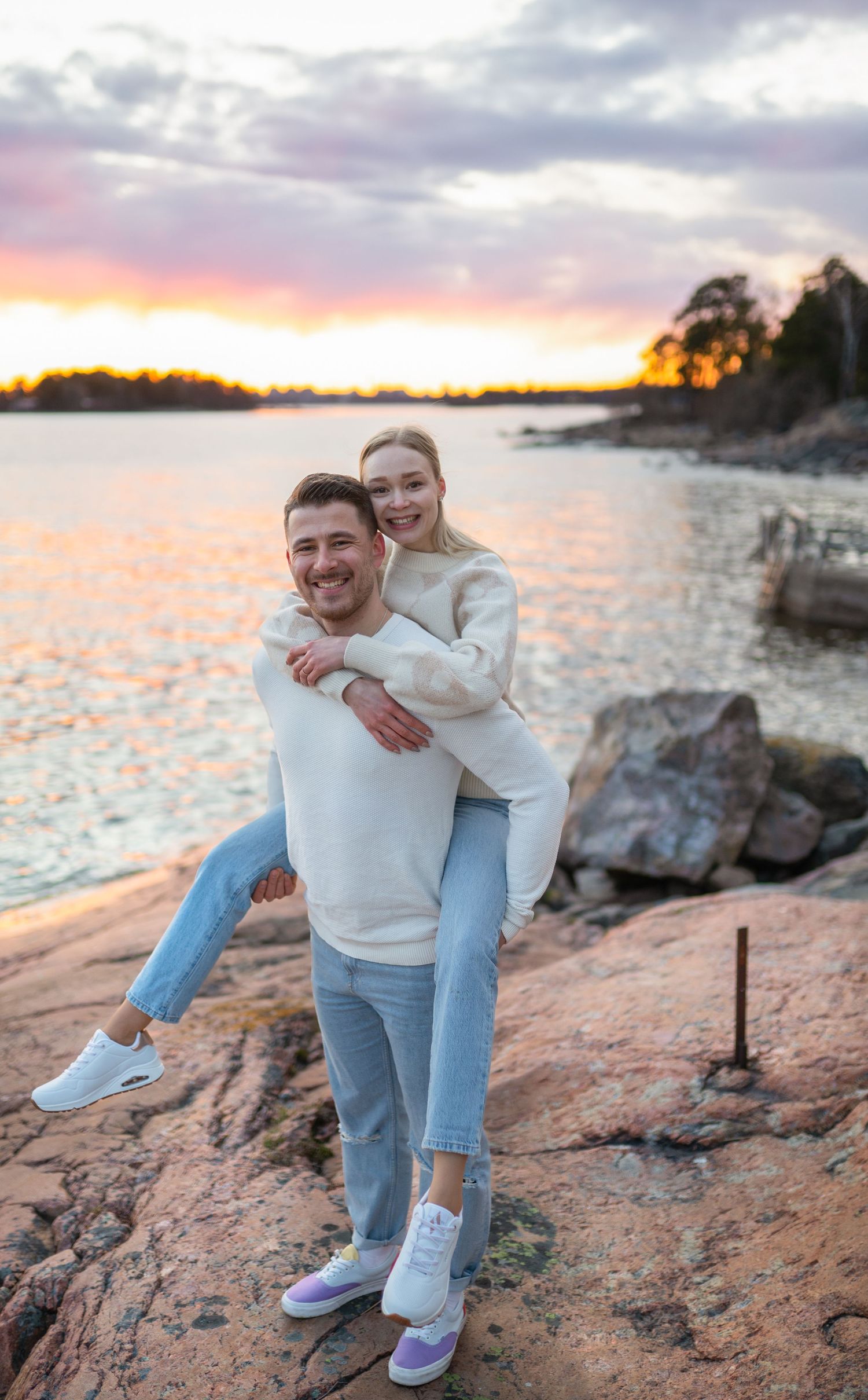 Man giving woman a piggyback ride on rocky lakeside at sunset with colorful sky reflection.