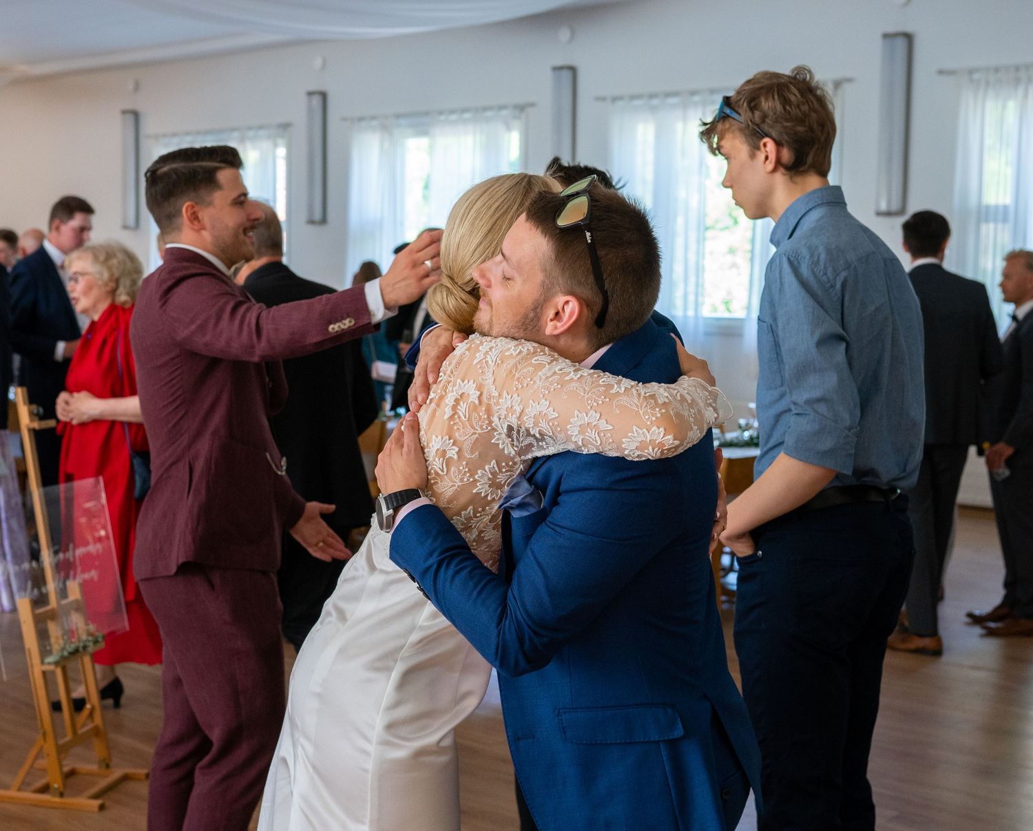 Bride in lace gown embraces male guest in blue suit at bright wedding reception venue.