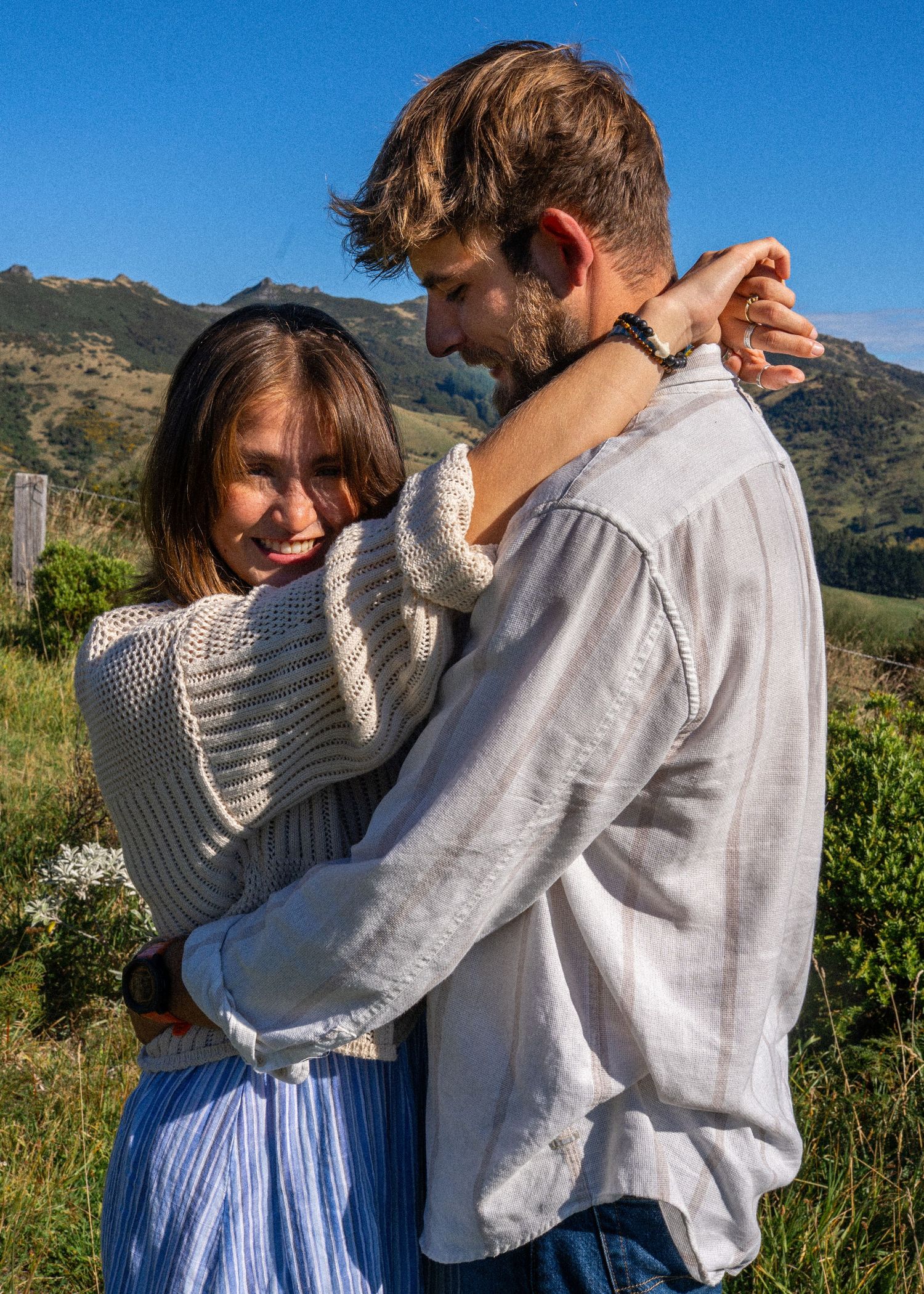 A happy couple embraces warmly in a lush green landscape with rolling hills and blue sky in the background.