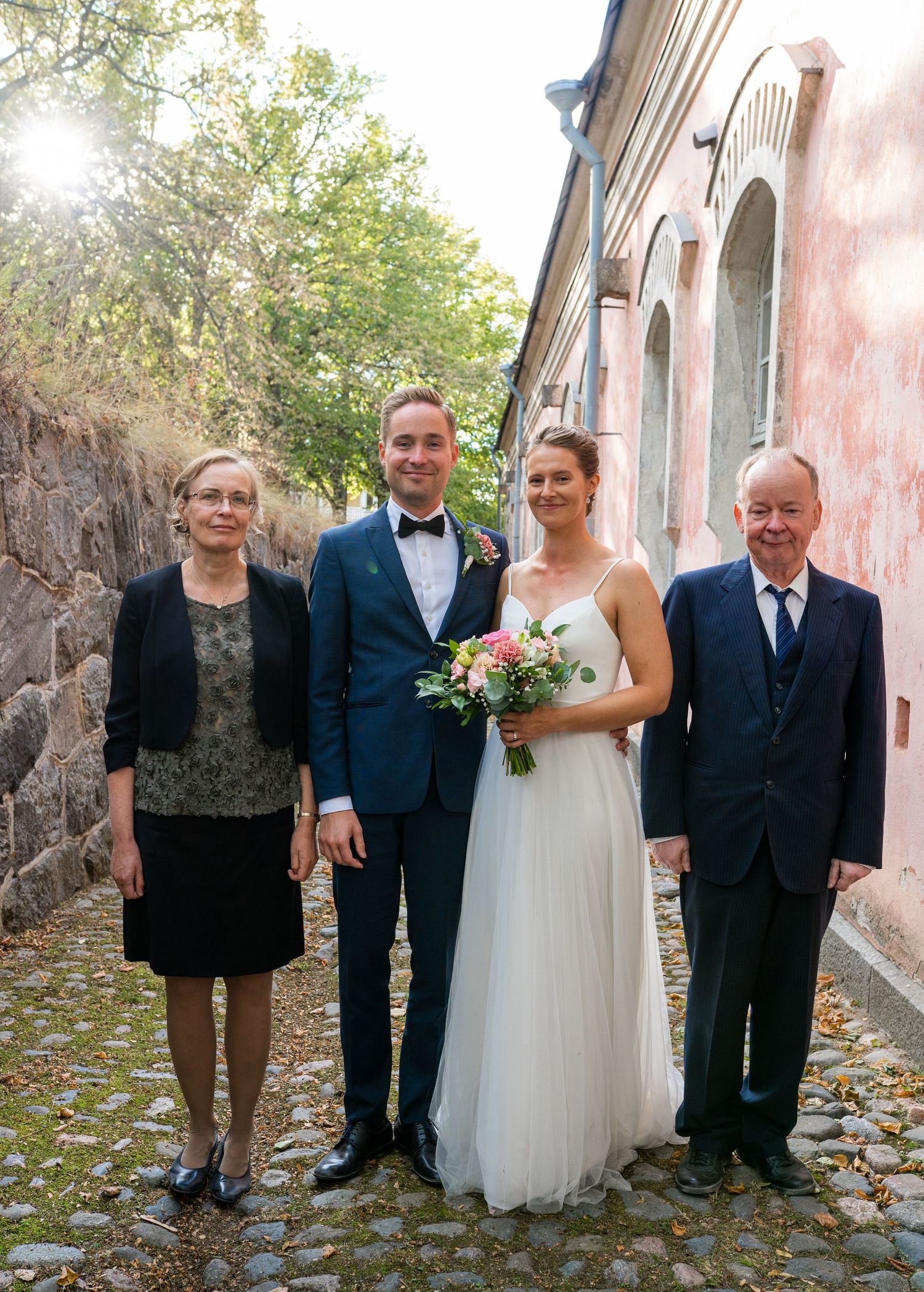 Bride and groom pose with two elderly relatives on cobblestone path beside pink building on wedding day.