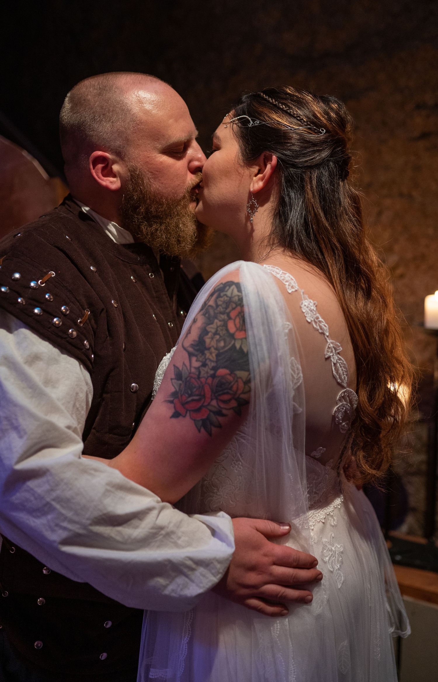 A couple shares a romantic wedding kiss, bride in lace gown with floral tattoo, groom in medieval-style brown vest.