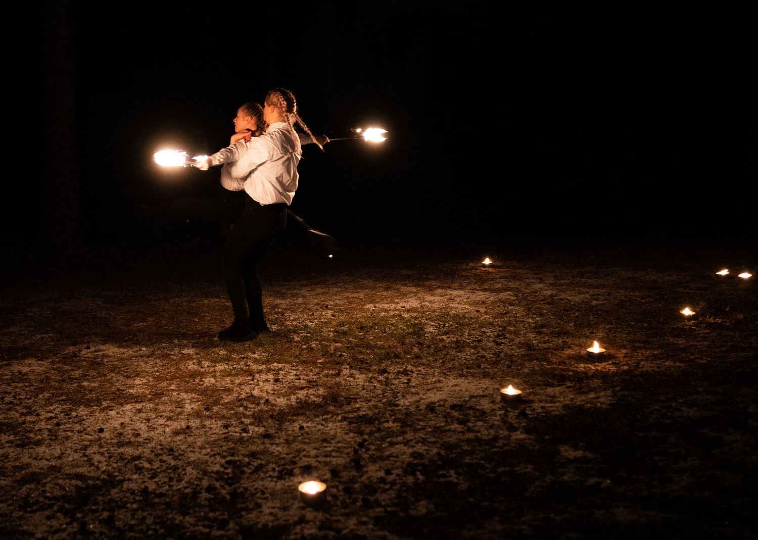 Person performing fire dancing at night, spinning glowing lights in a dark outdoor setting with candles on the ground.