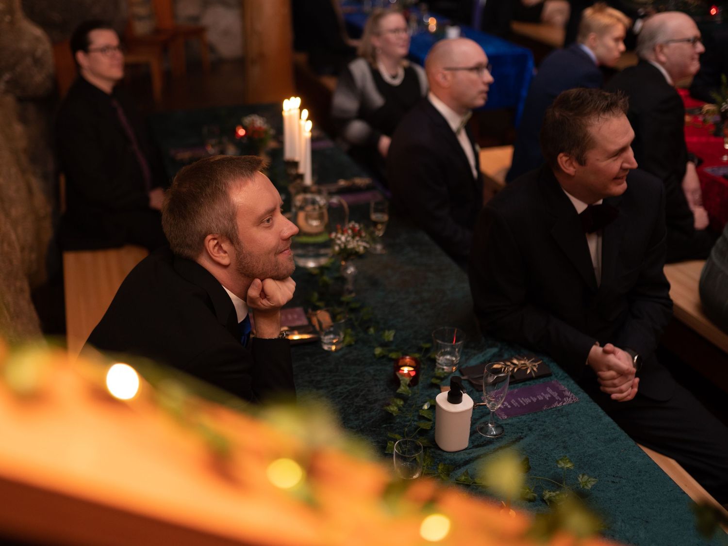 Two men in tuxedos sit at a candlelit table during a formal evening event in an elegant venue.