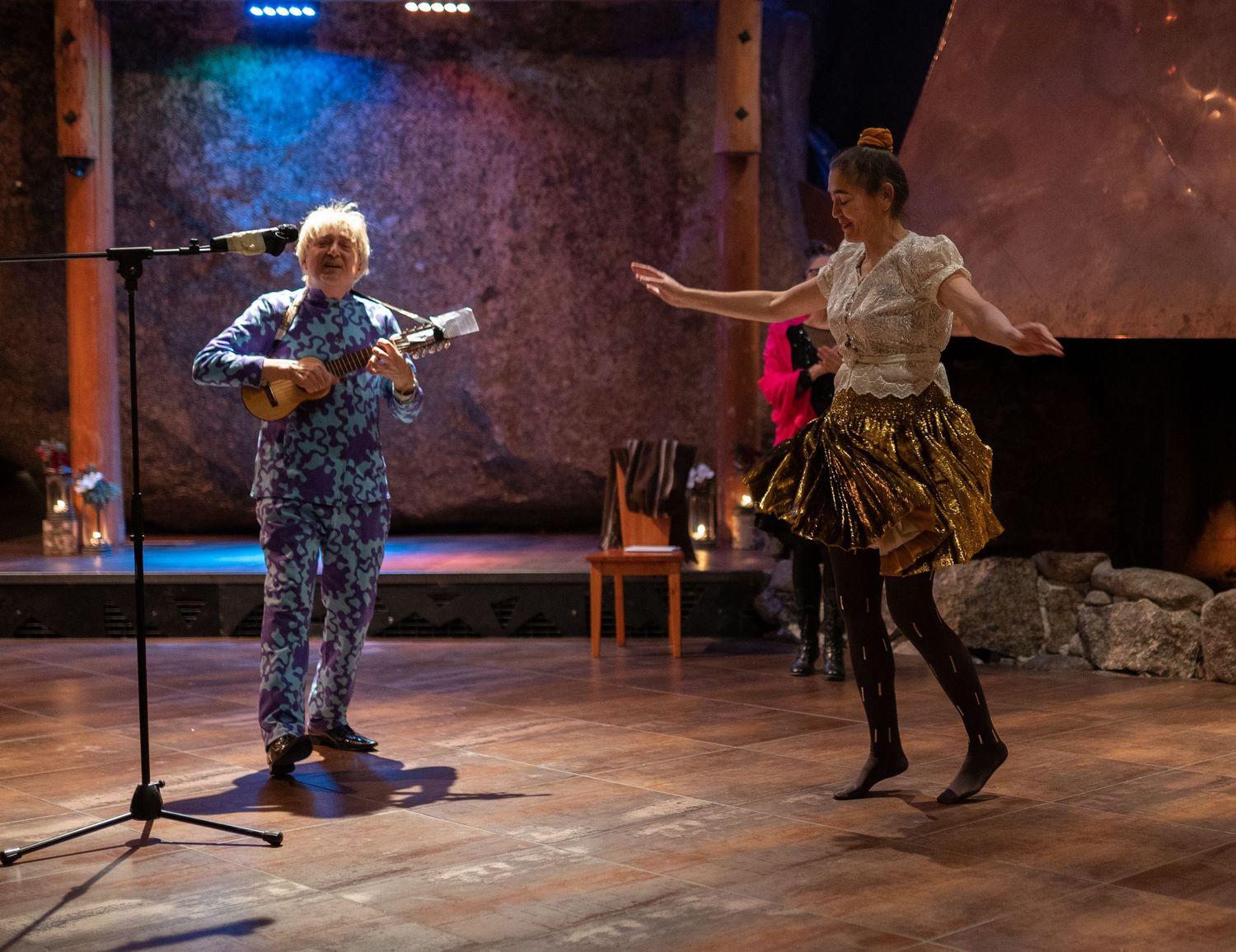 Musician in floral suit plays ukulele at mic while dancer in gold skirt performs on stage.