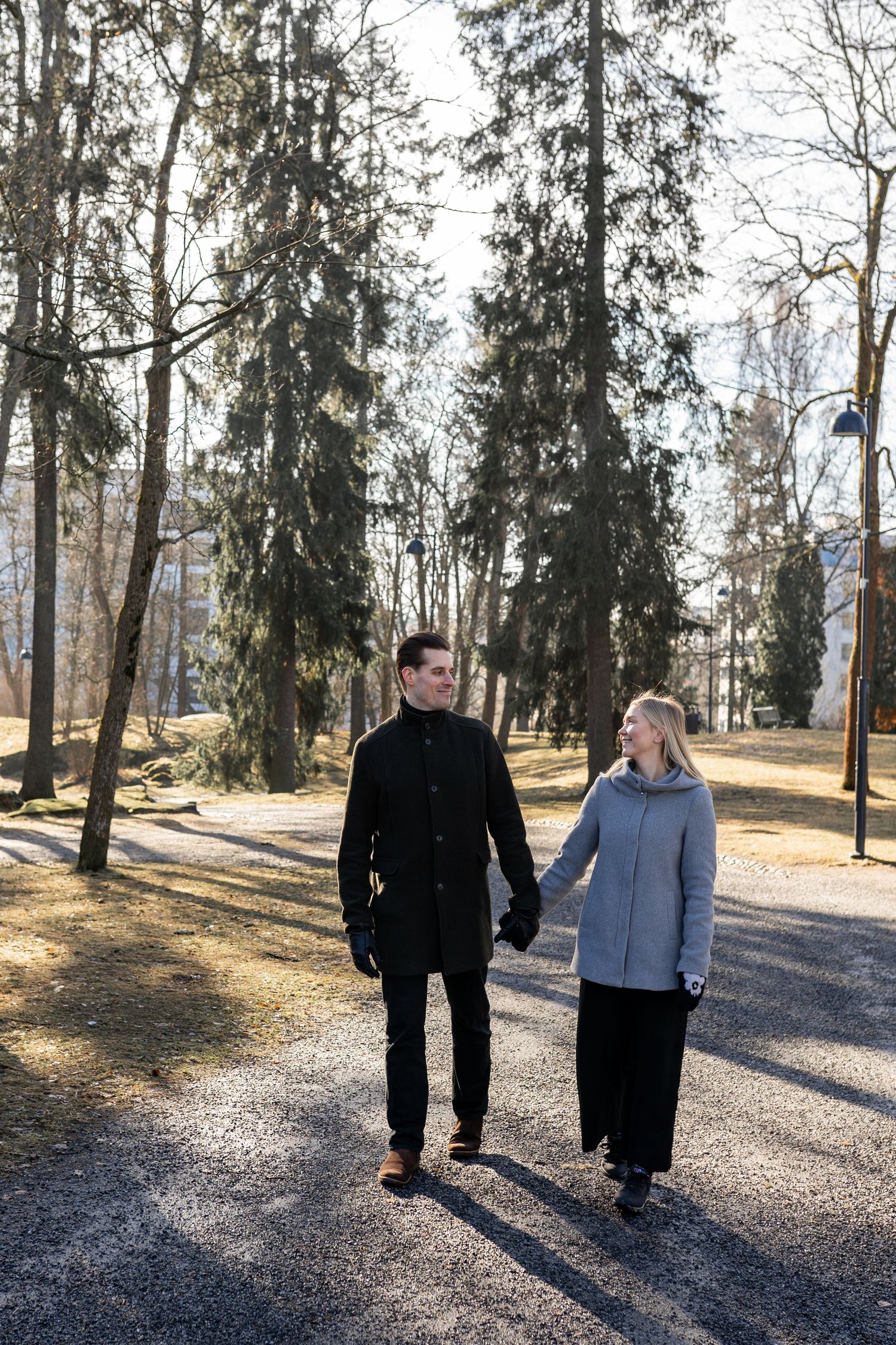 Couple holding hands walking through a sunlit park in winter coats among tall trees.