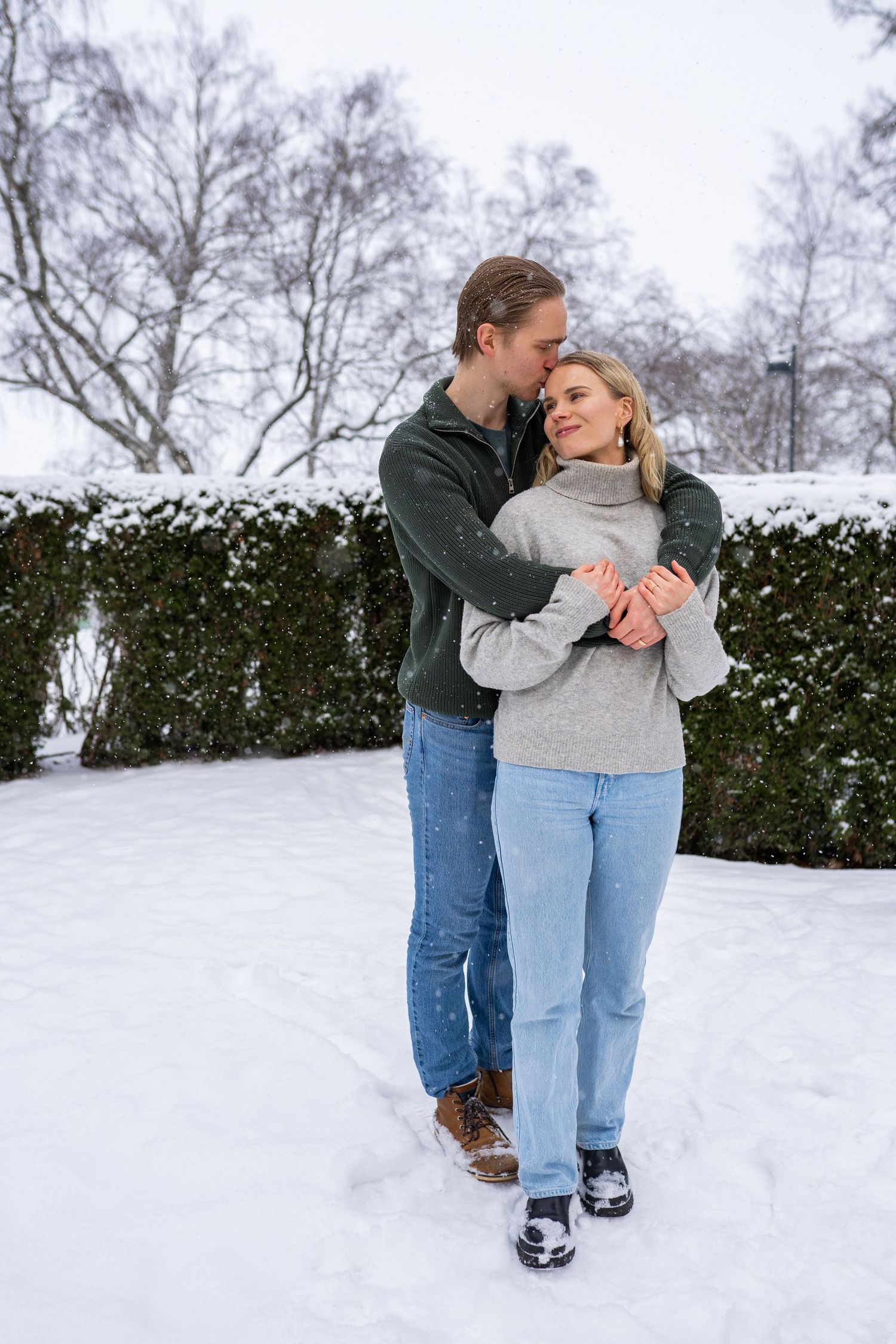 Couple embracing in snowy winter park, man in green jacket hugging woman in gray sweater and jeans.
