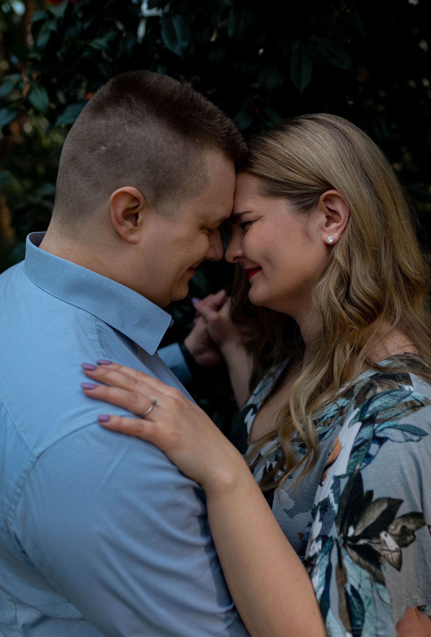 A couple shares a romantic forehead-to-forehead moment outdoors, the woman wearing a floral dress and engagement ring.