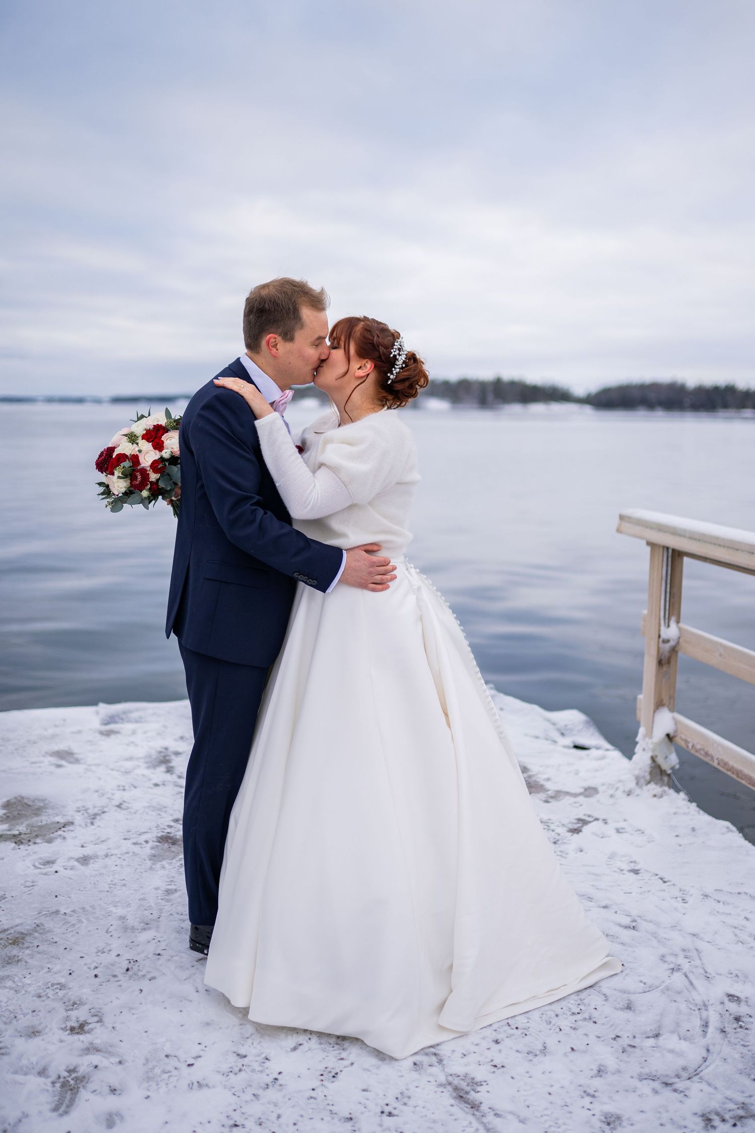 Bride and groom share a kiss on a snowy winter pier, she in white gown, he in navy suit with red bouquet.