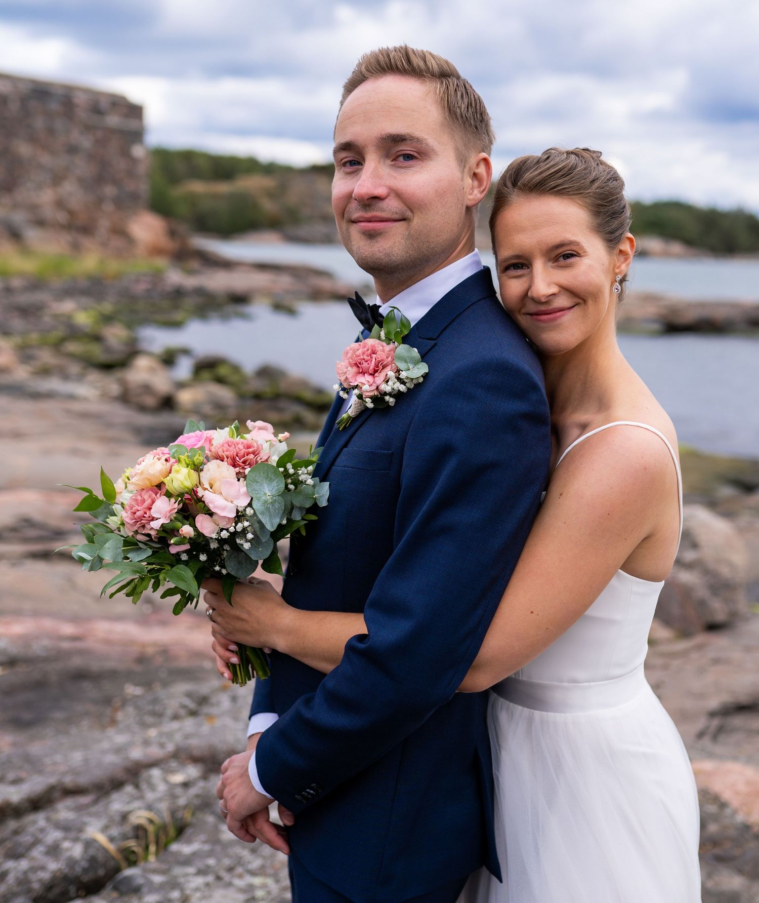 Wedding couple poses by rocky seaside, bride holding pink bouquet, groom in navy suit.