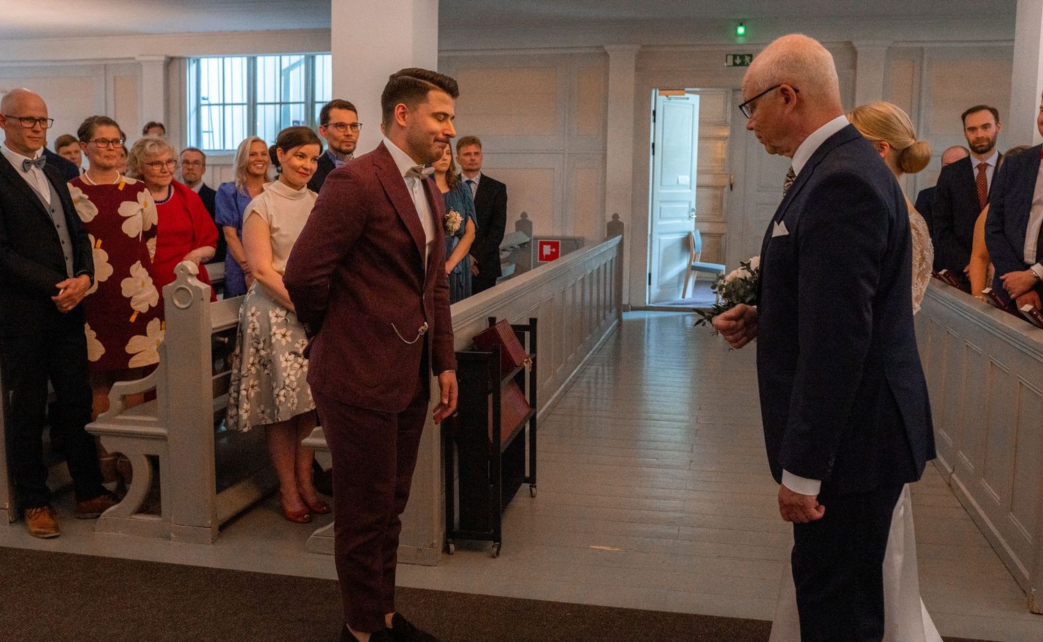 Groom in burgundy suit greeting man in navy suit at church altar during wedding ceremony.