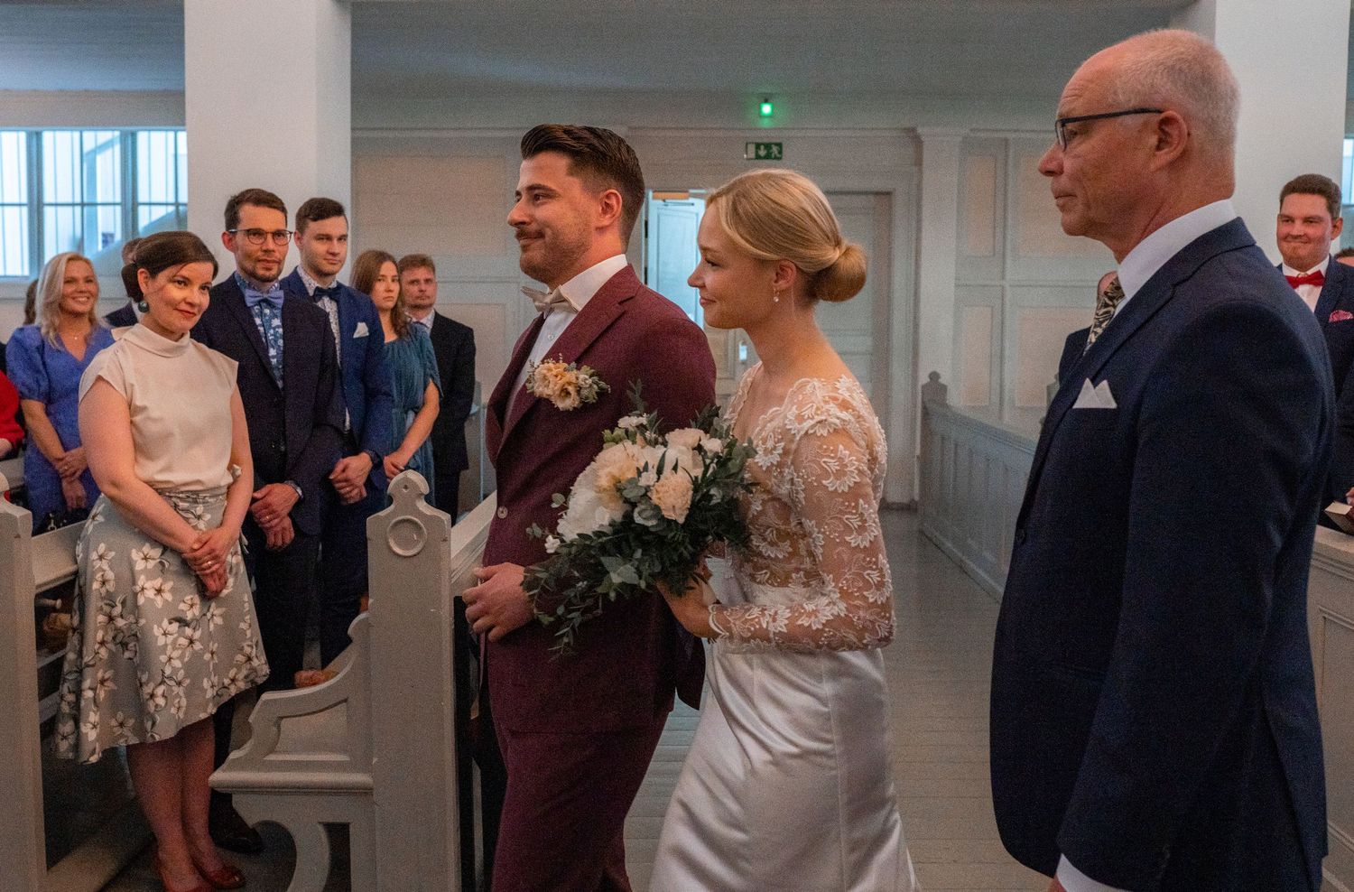 Smiling bride holding bouquet stands with groom in burgundy suit during church wedding ceremony with guests.