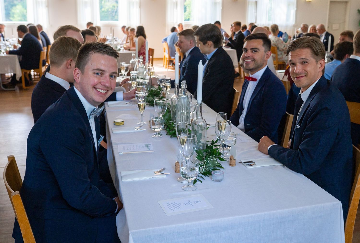 Four young men in navy suits smiling at wedding reception dinner table.
