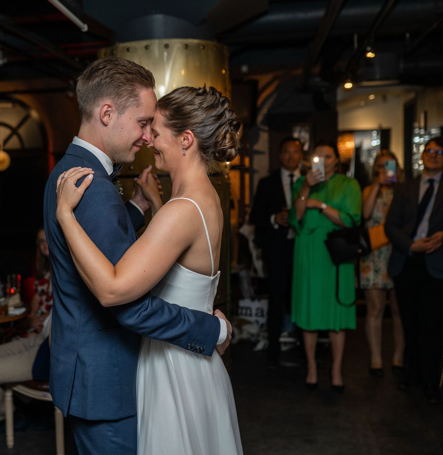 Bride and groom share a tender moment during first dance, nearly touching foreheads, guests watching in background.