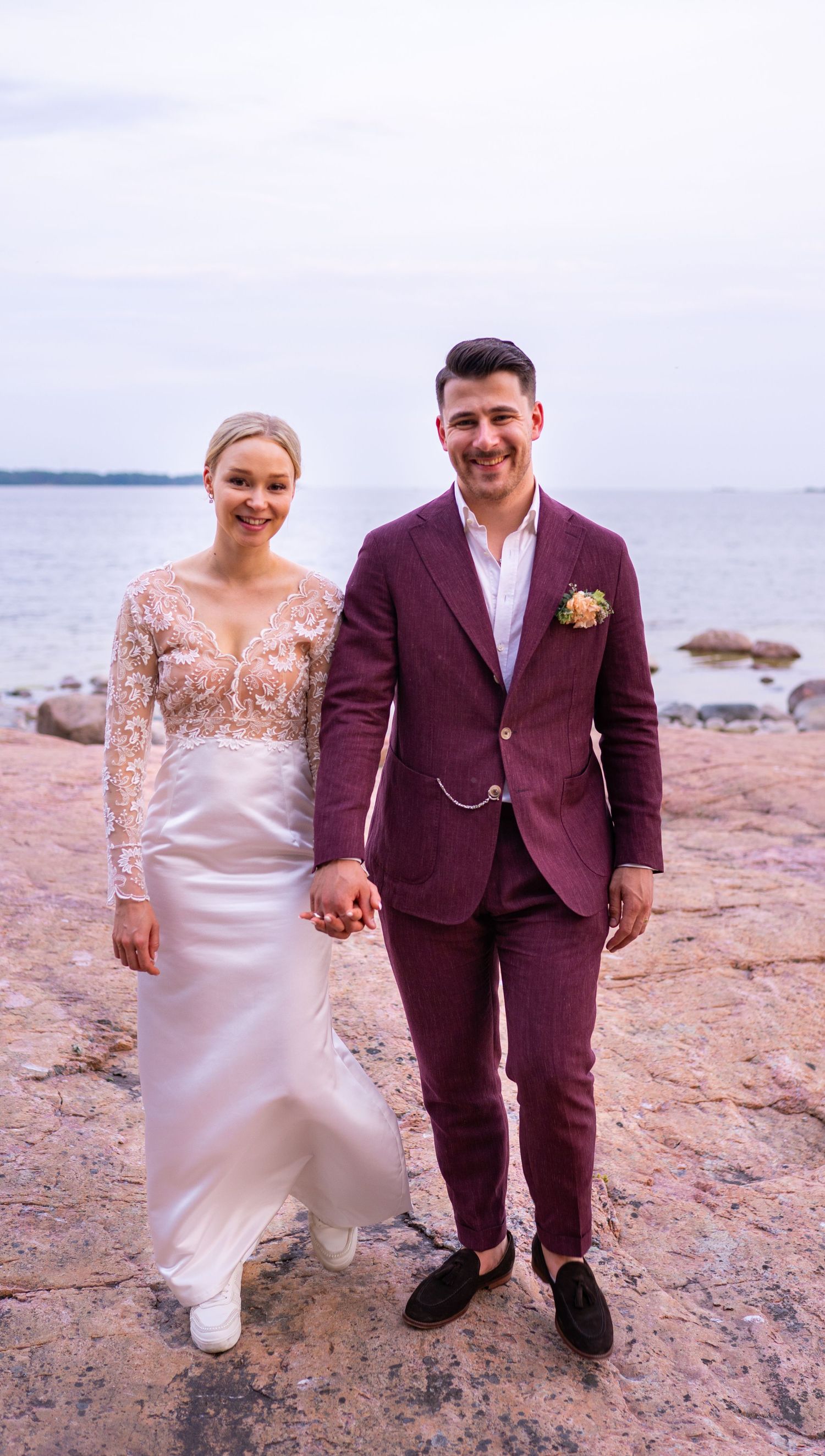 Bride and groom holding hands and smiling on rocky shore, bride in lace gown and groom in burgundy suit.