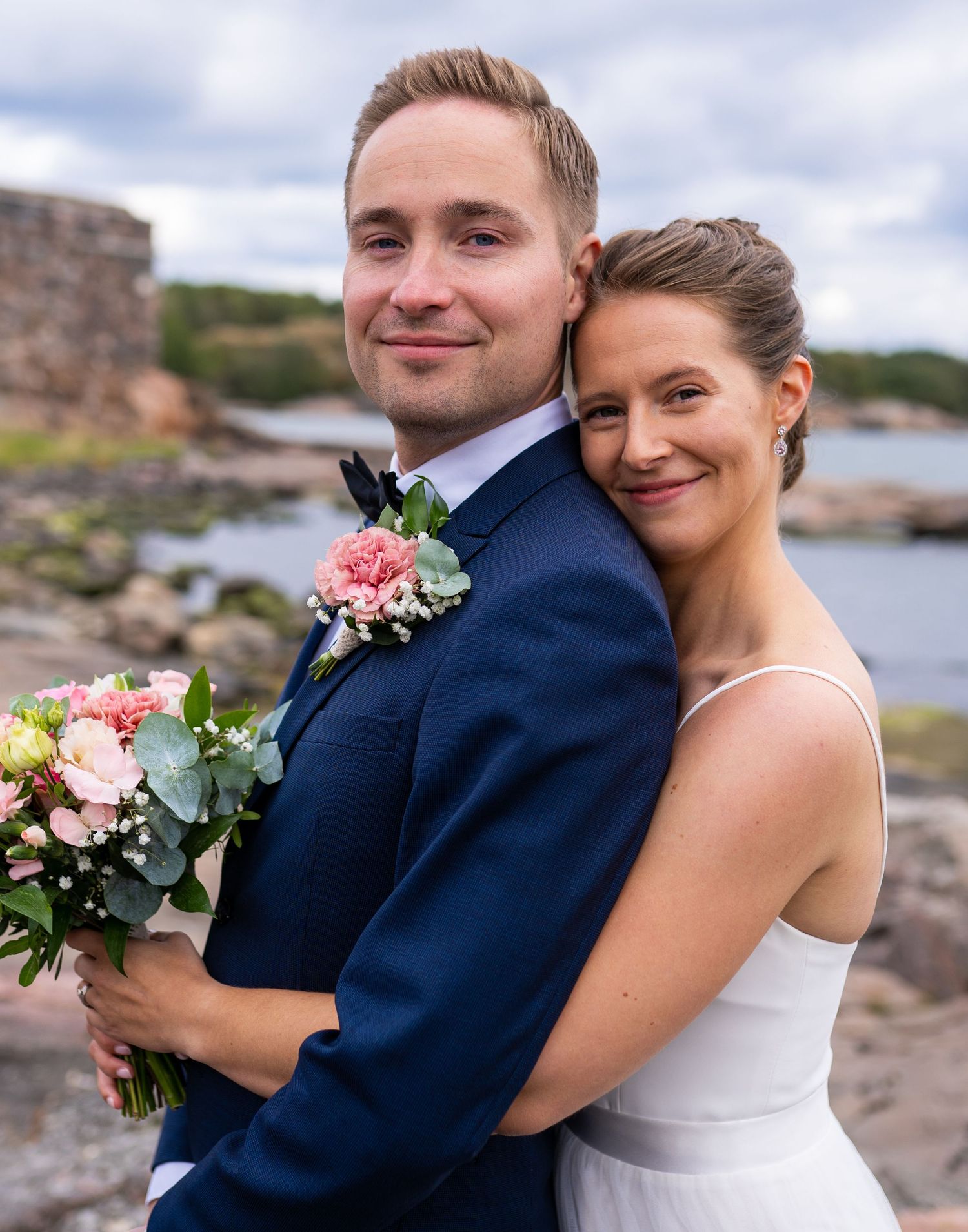 Bride embracing groom from behind on rocky coast, both smiling, she holds pink floral bouquet, groom in navy suit.