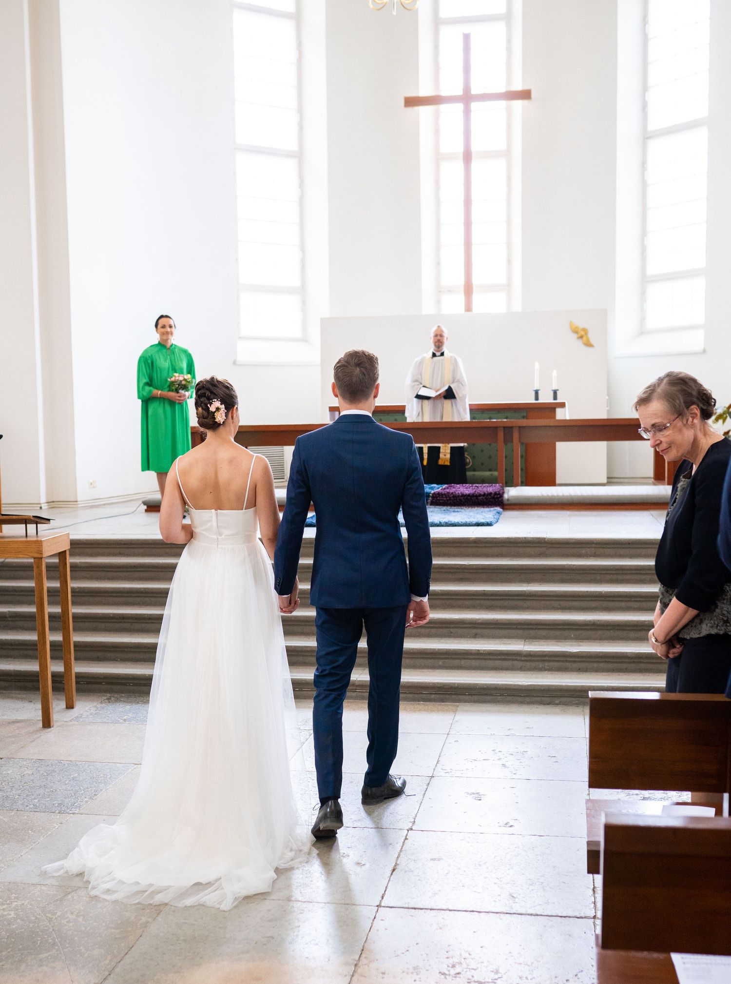 Bride and groom walking toward the altar where a pastor and woman in green dress await.