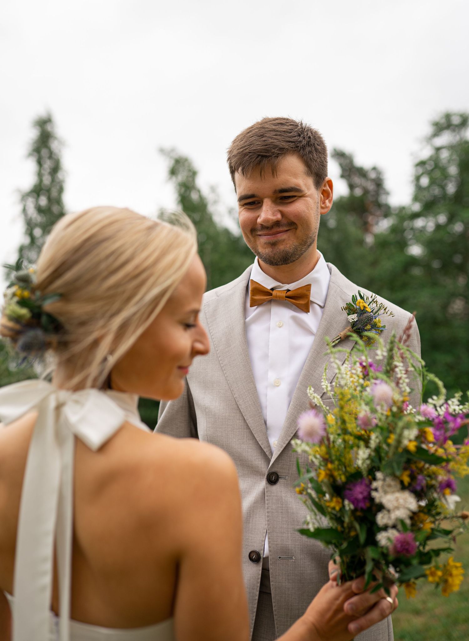 Groom in grey suit with brown bow tie smiling at bride holding colorful wildflower bouquet outdoors.