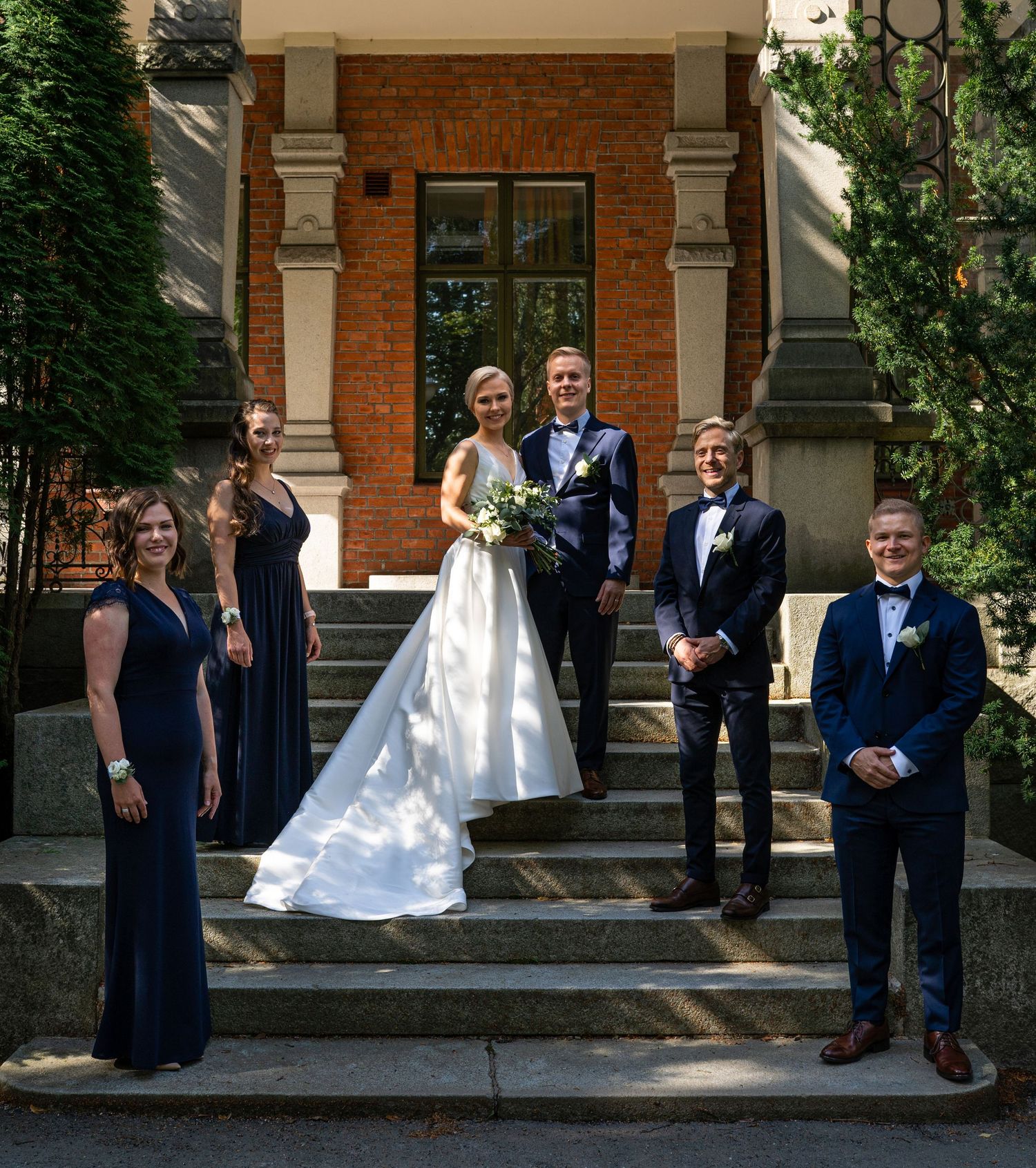 Wedding party posing on stone steps outside brick building, bride in white gown with groomsmen and bridesmaids.
