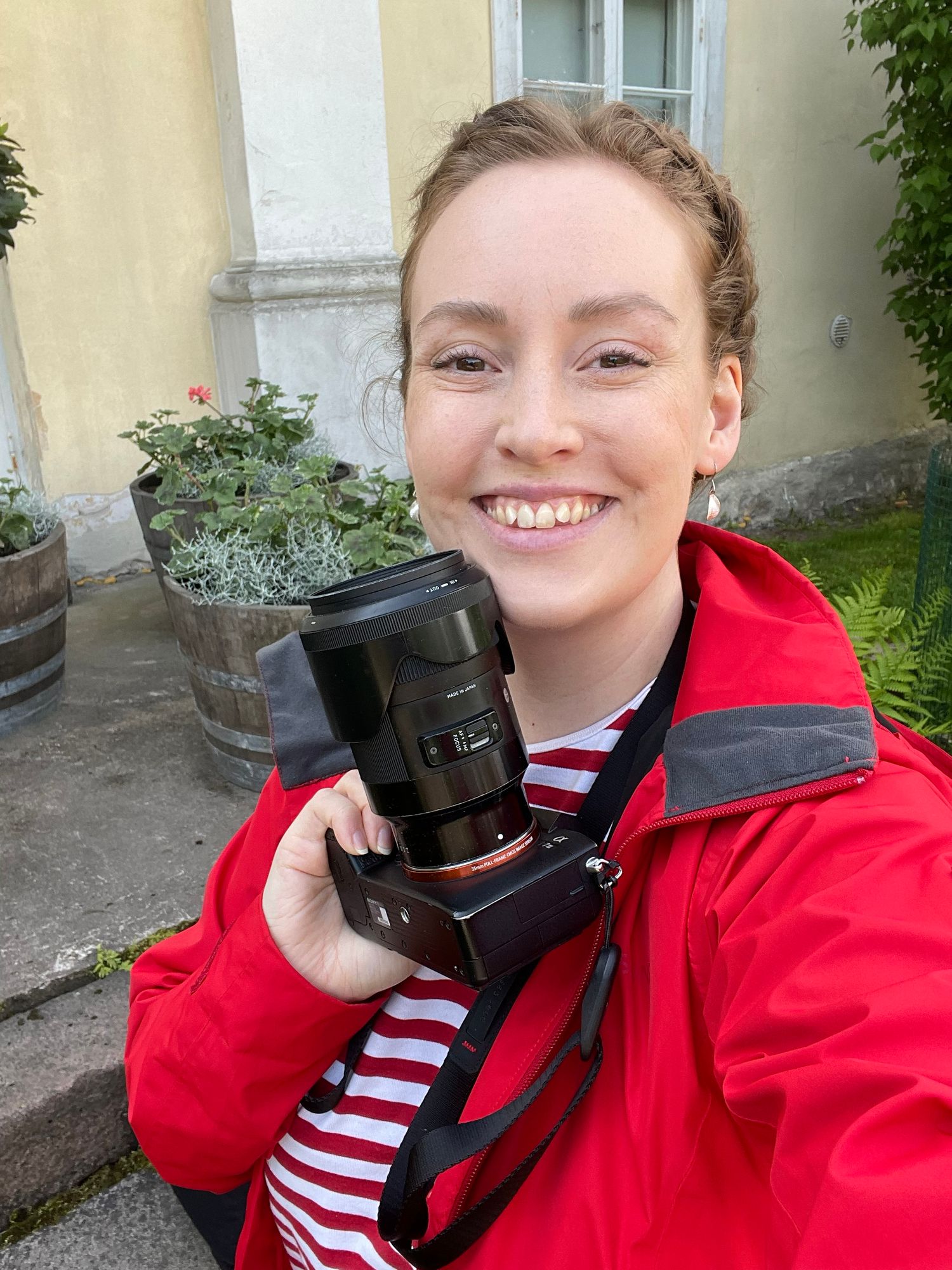 Smiling young woman in red jacket holds a professional camera outdoors near a yellow building.