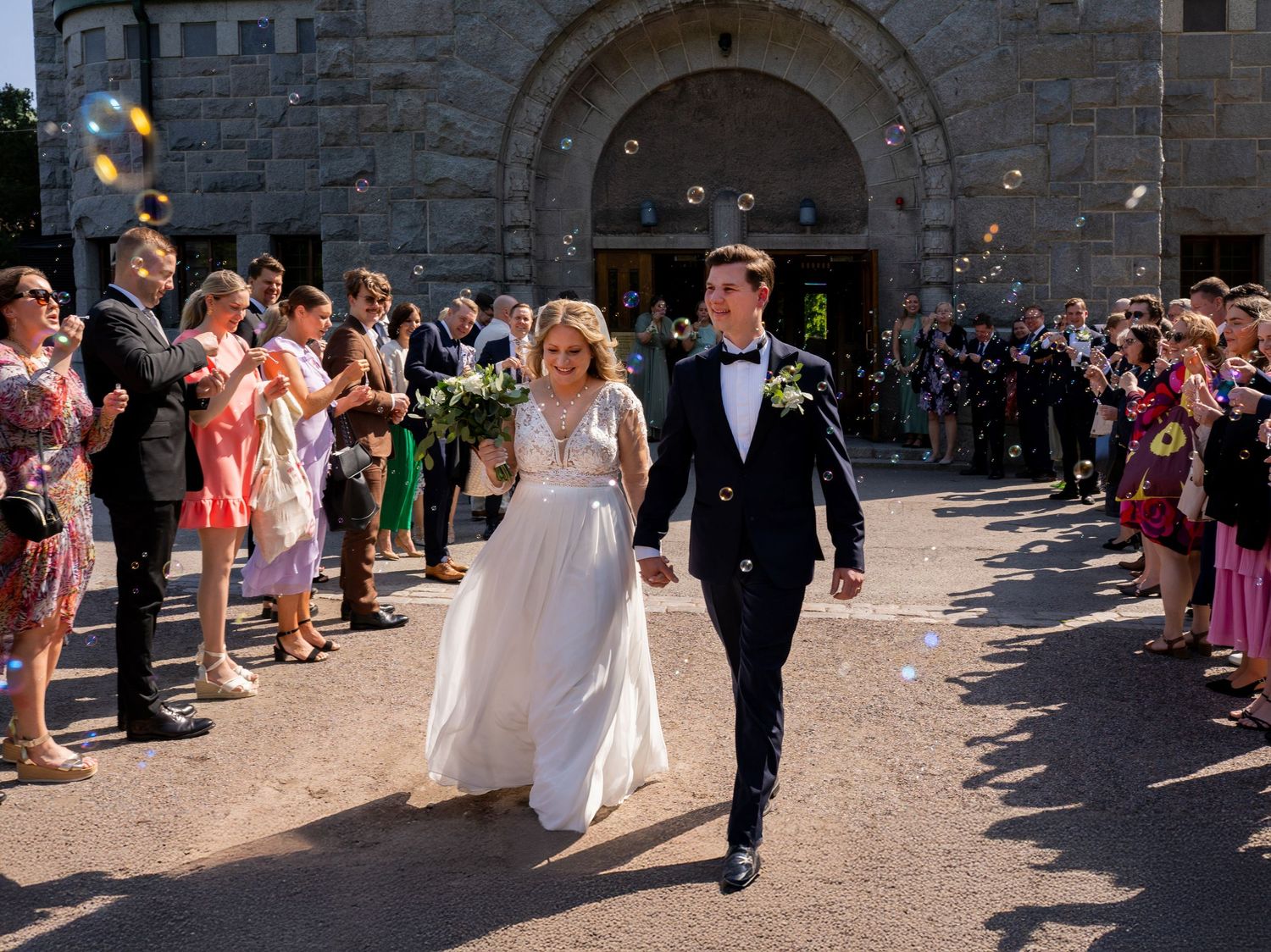 Newlyweds walking through cheering guests blowing bubbles outside stone church after their wedding ceremony.