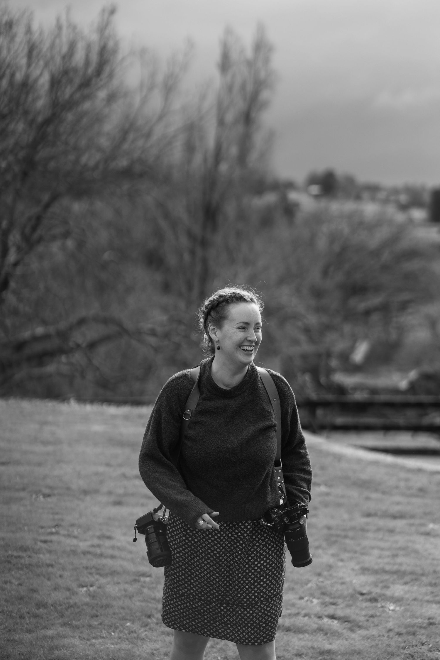 Black and white photo of a smiling woman holding cameras, wearing a skirt and sweater, standing outdoors near bare trees.