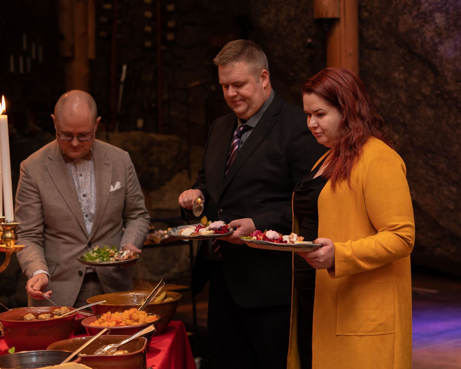 Three people serving themselves at a buffet with colorful dishes in a warmly lit dining venue.