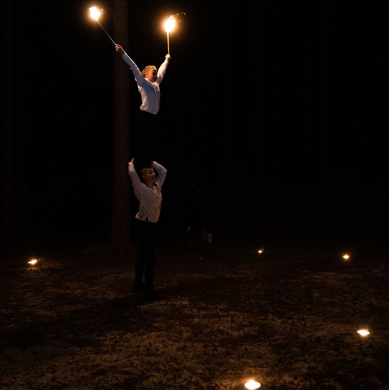Two performers doing an acrobatic act with fire torches at night, surrounded by glowing lights on the ground.