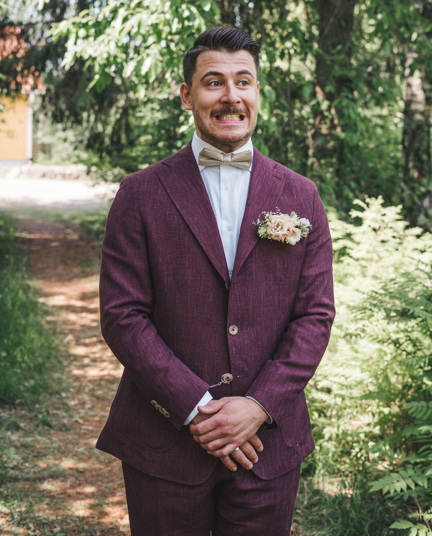 Smiling groom in burgundy tweed suit with floral boutonniere and beige bow tie in forest.