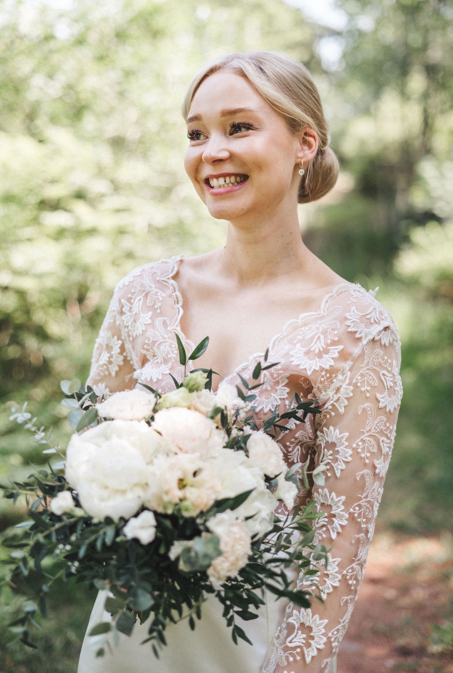 Joyful bride in lace wedding dress holding white peony bouquet in sunlit forest.