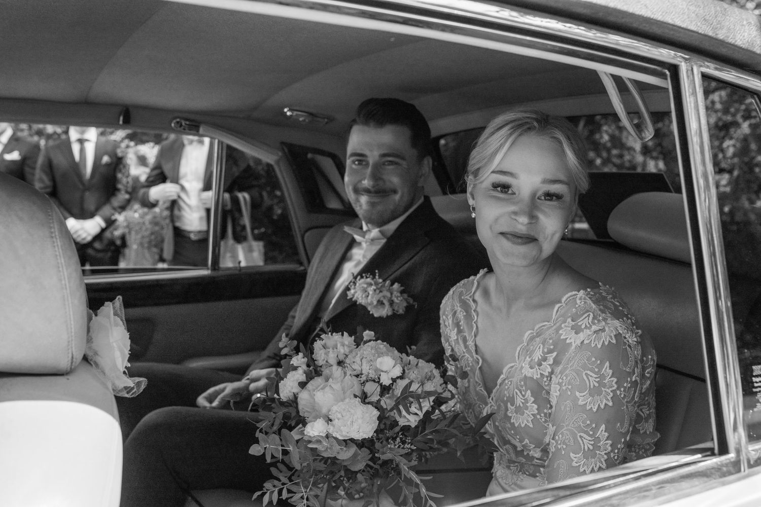 Black and white portrait of smiling bride and groom seated together inside vintage wedding car.