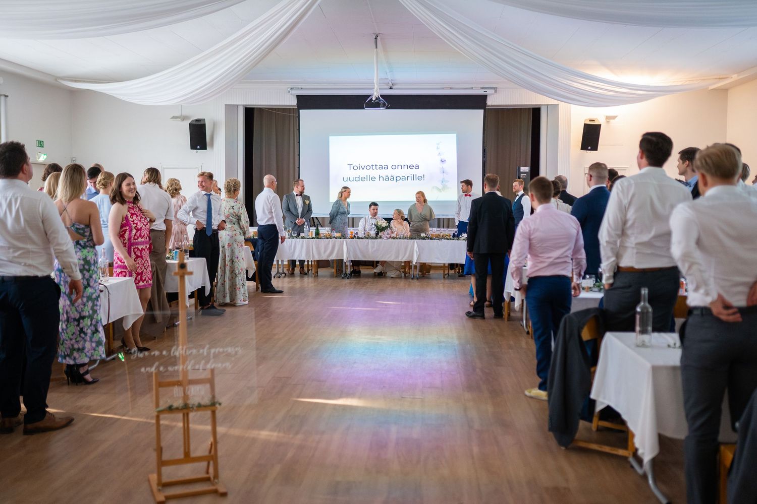 Guests mingle in a bright wedding reception hall with a projection screen and colorful light reflections.