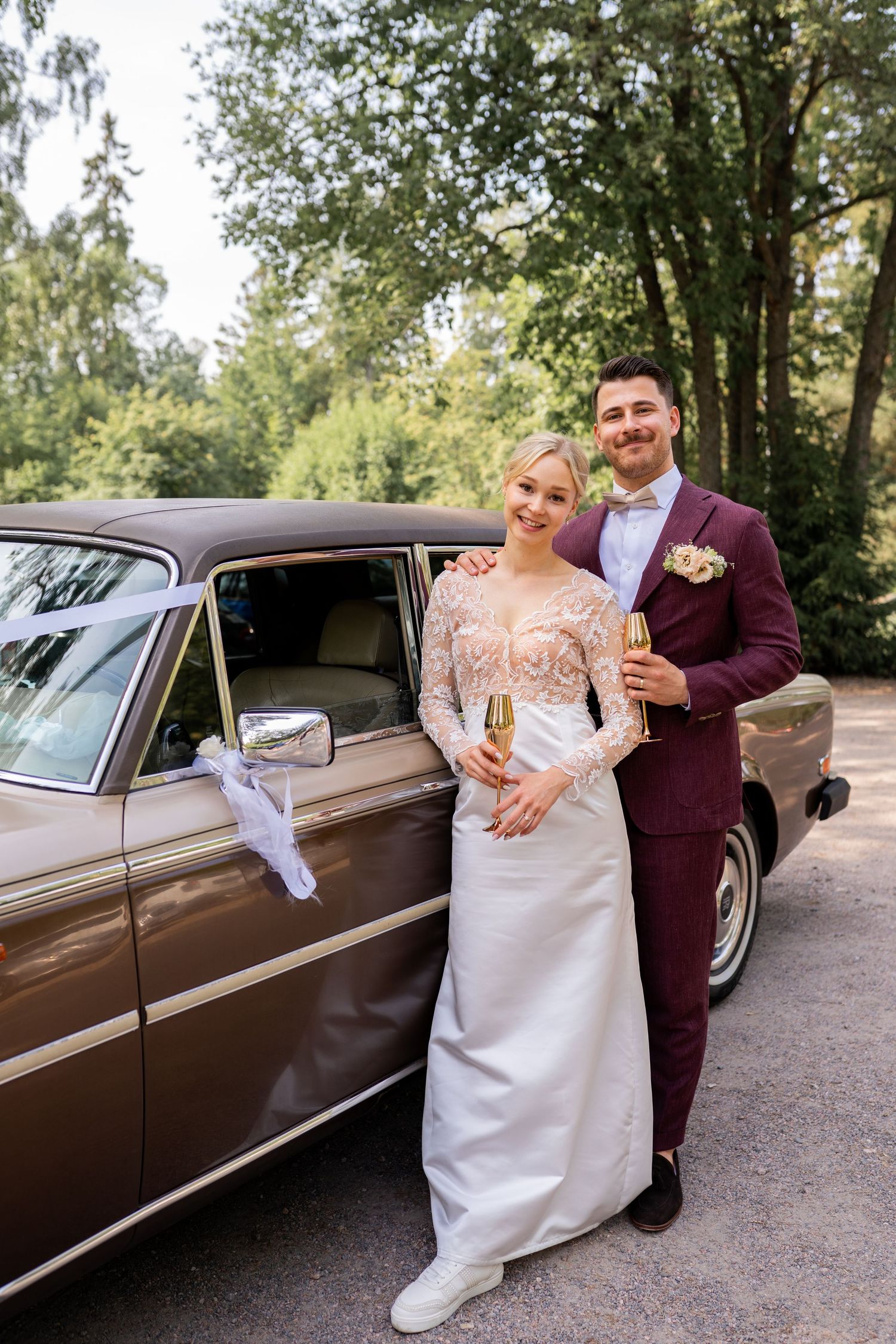 Bride and groom holding champagne flutes beside a vintage brown car on their wedding day.