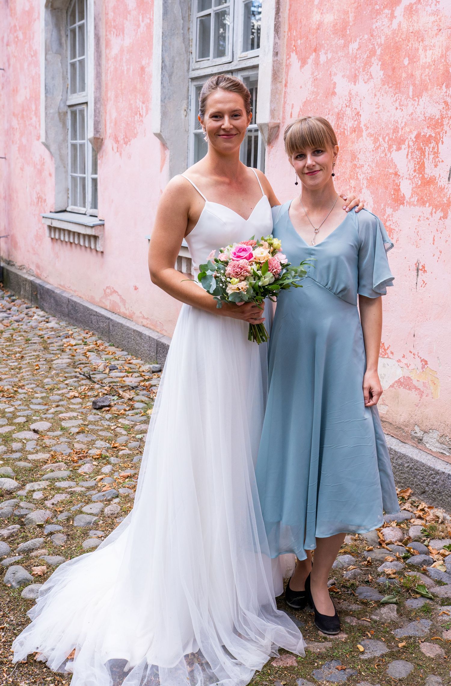 Tall bride in white spaghetti-strap gown with pink bouquet poses with guest in light blue midi dress.