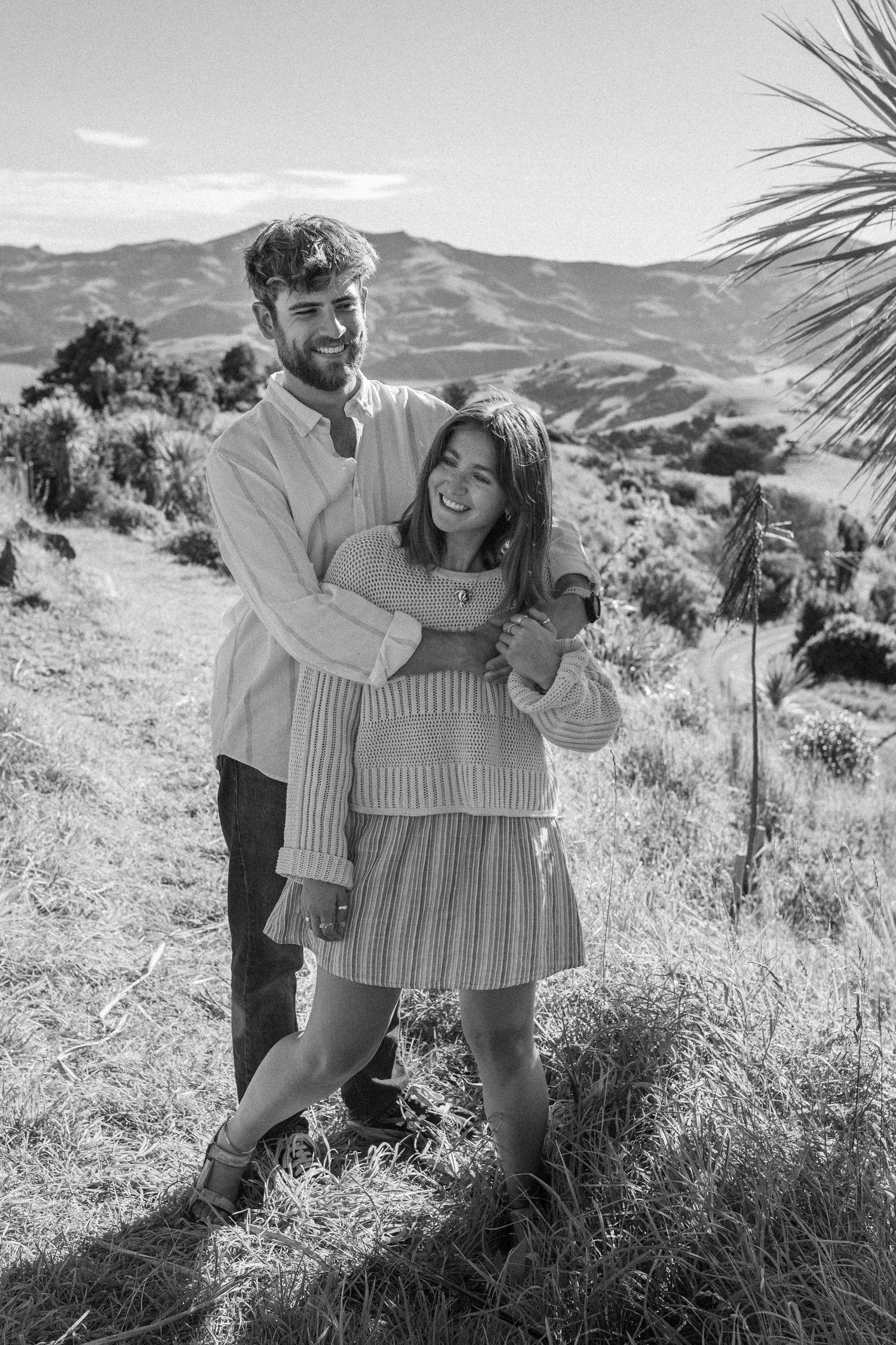 A joyful couple embraces and laughs together in a black and white photo on a sunny hillside.
