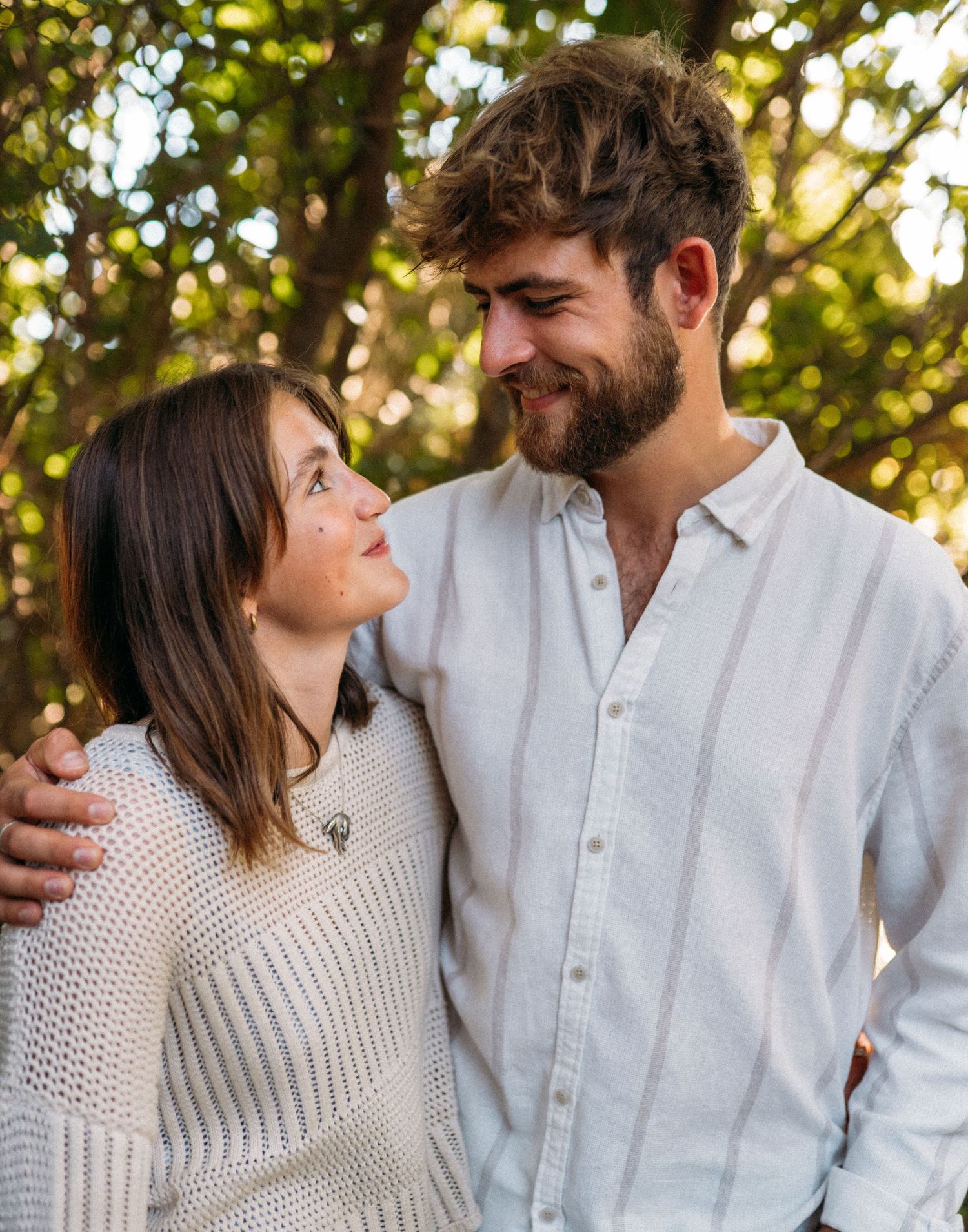 Happy couple smiling at each other with arms around shoulders in a sunny park.