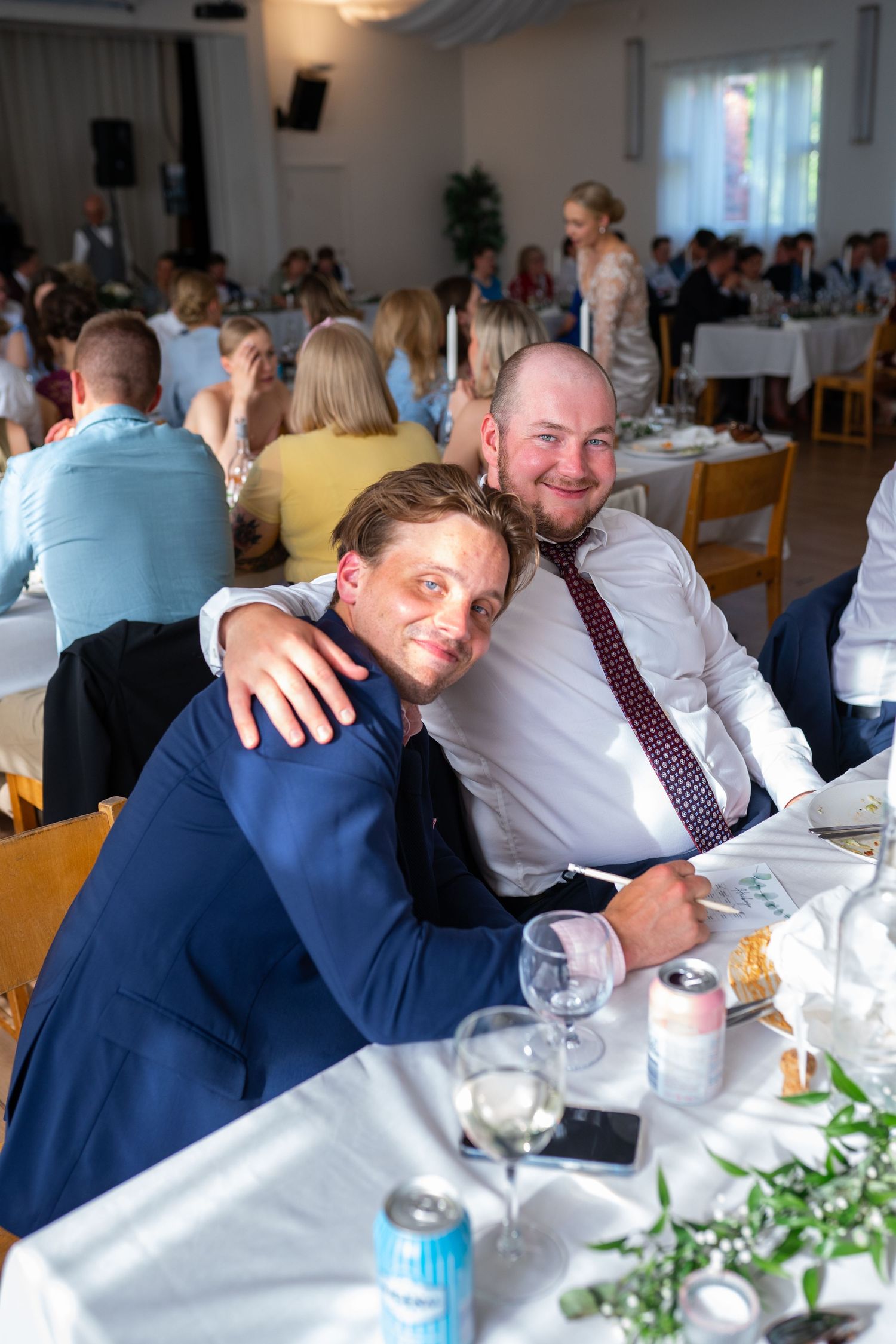 Two smiling men in formal attire posing together at a wedding reception dinner table.