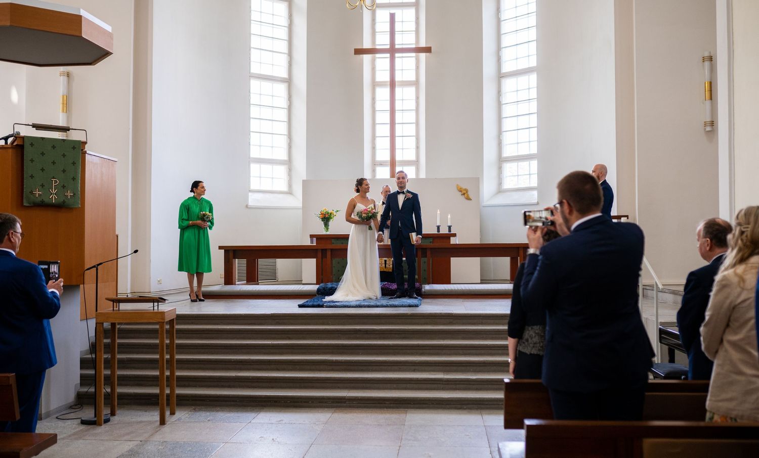 Couple standing at church altar while guests photograph the wedding ceremony.