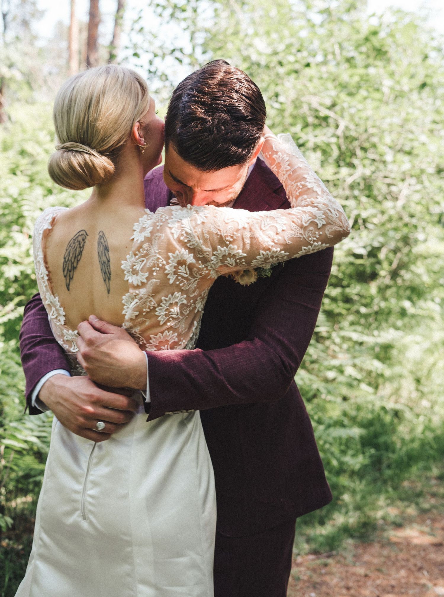 Groom in burgundy suit kisses bride with angel wing tattoo on back during outdoor forest wedding embrace.