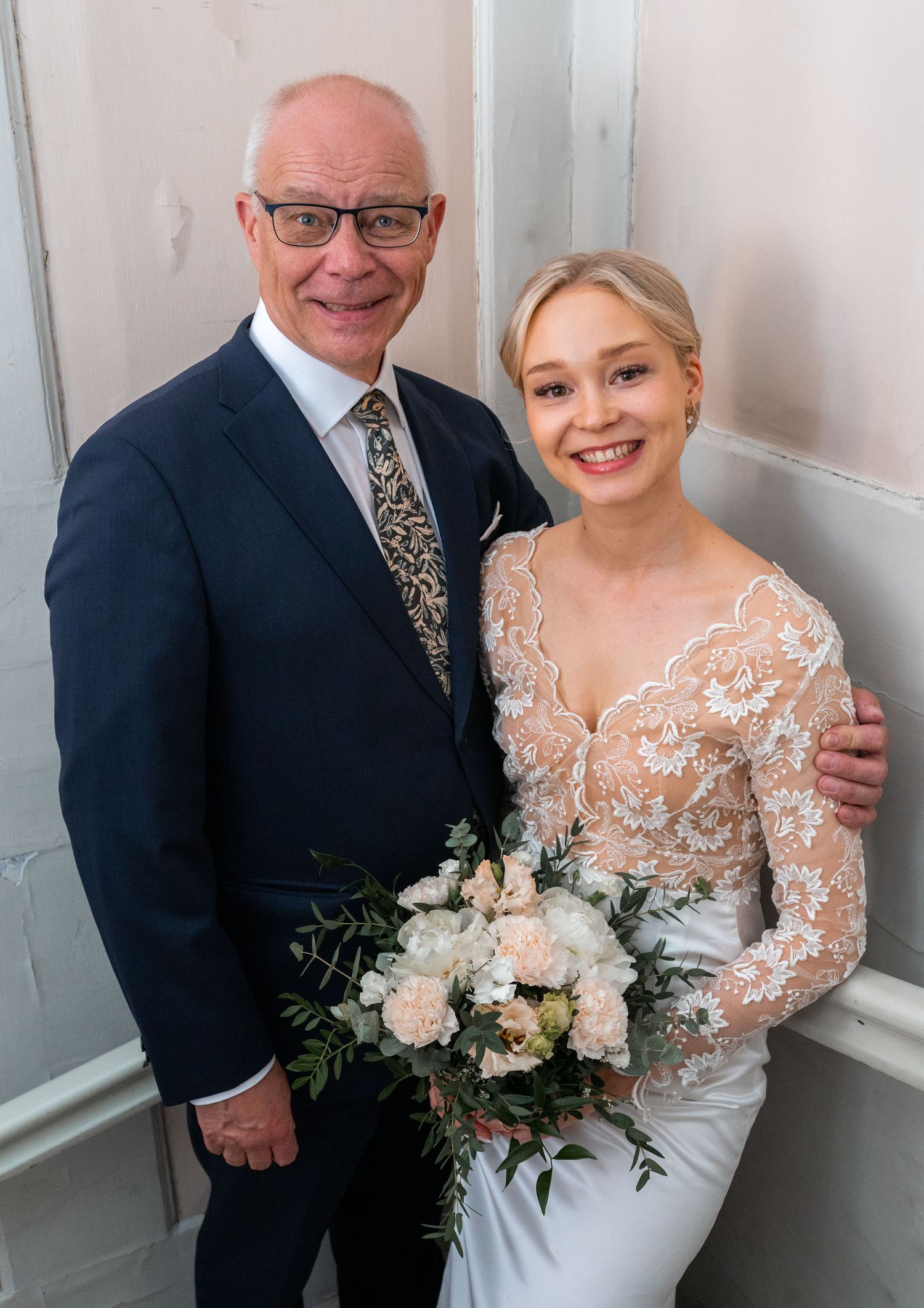 Smiling bride in lace wedding dress holding white bouquet posing with older man in navy suit.